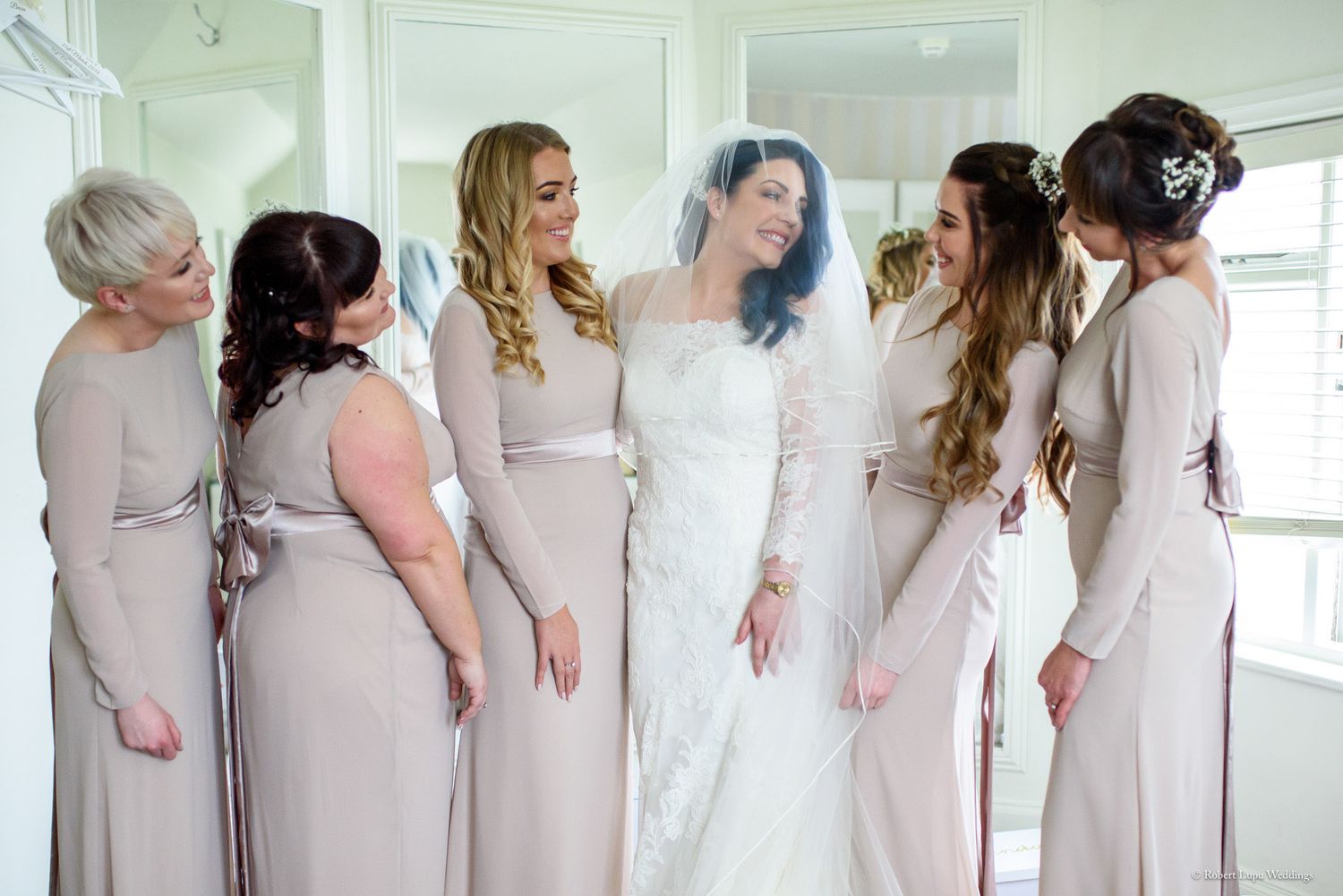 Bride in white lace gown with bridesmaids in taupe dresses smiling together before the wedding ceremony.
