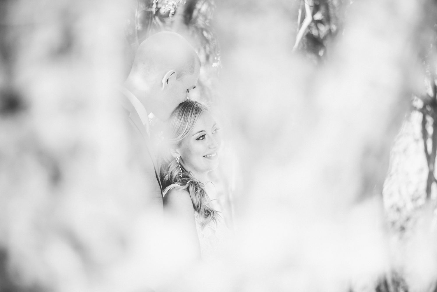 Dreamy black and white portrait of bride seen through soft-focus foliage in nature setting.
