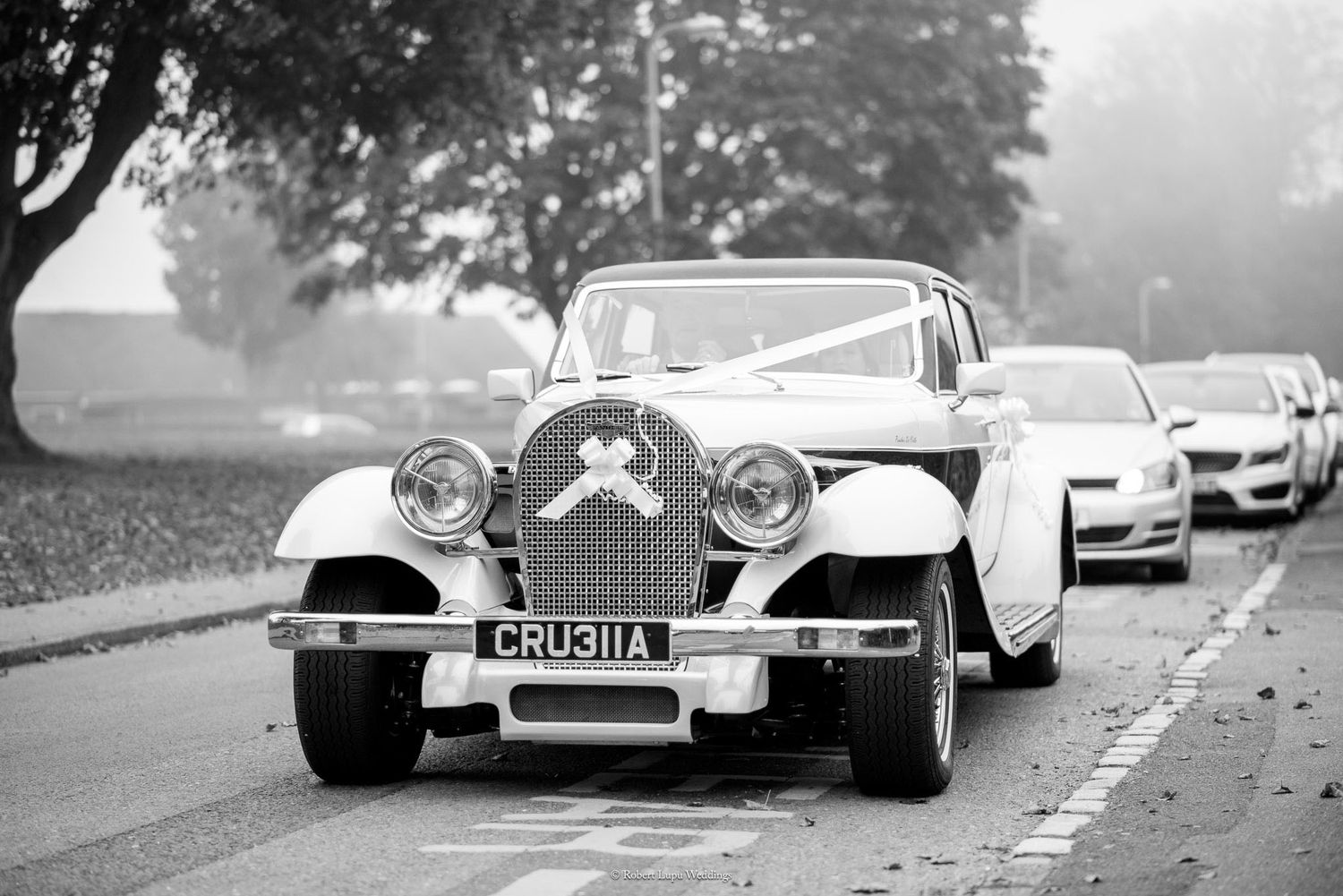 Vintage white wedding car decorated with ribbons leading a bridal procession on a misty road.