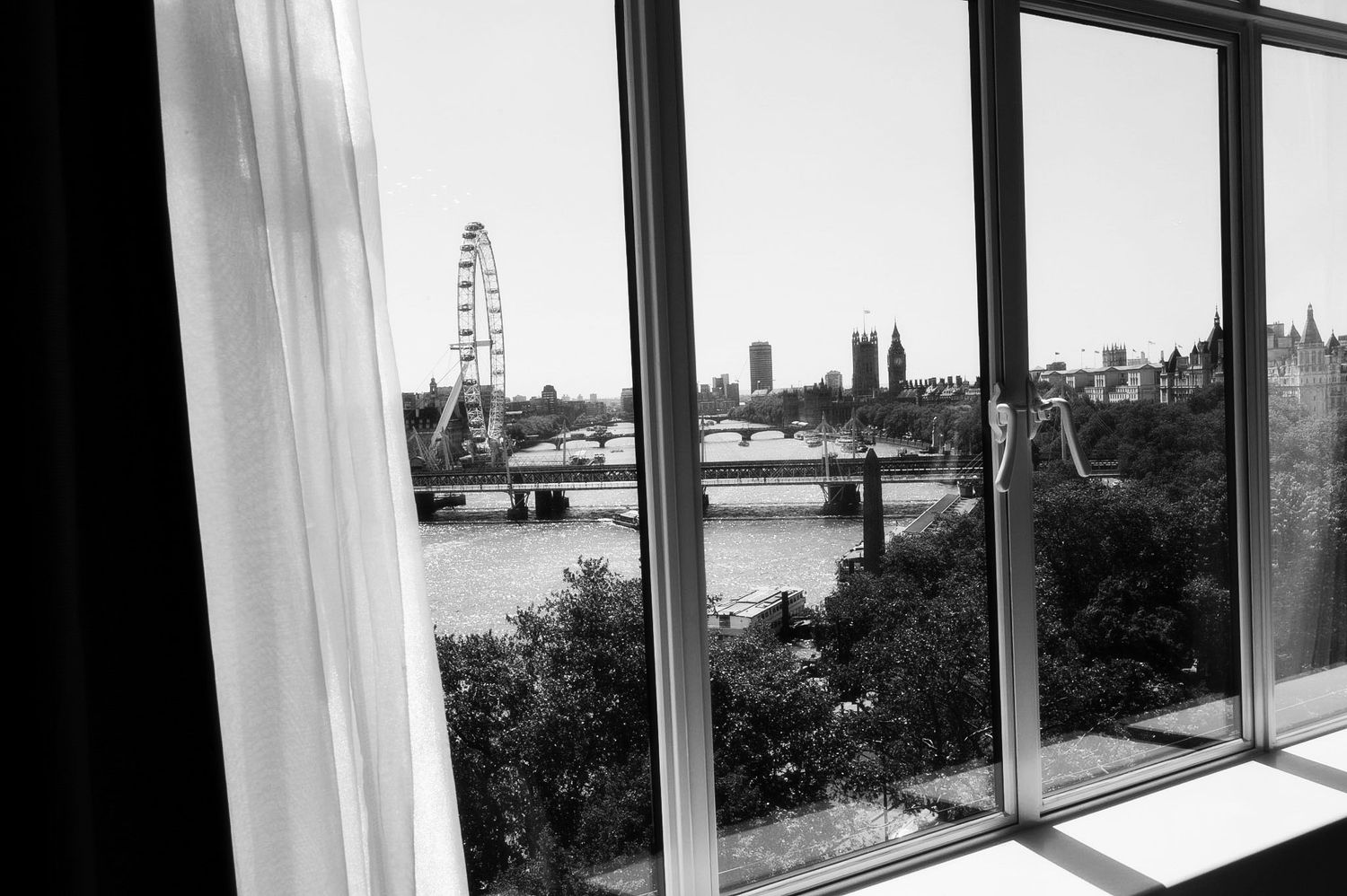 Black and white view of London Eye and Thames River from a hotel window with sheer curtains.