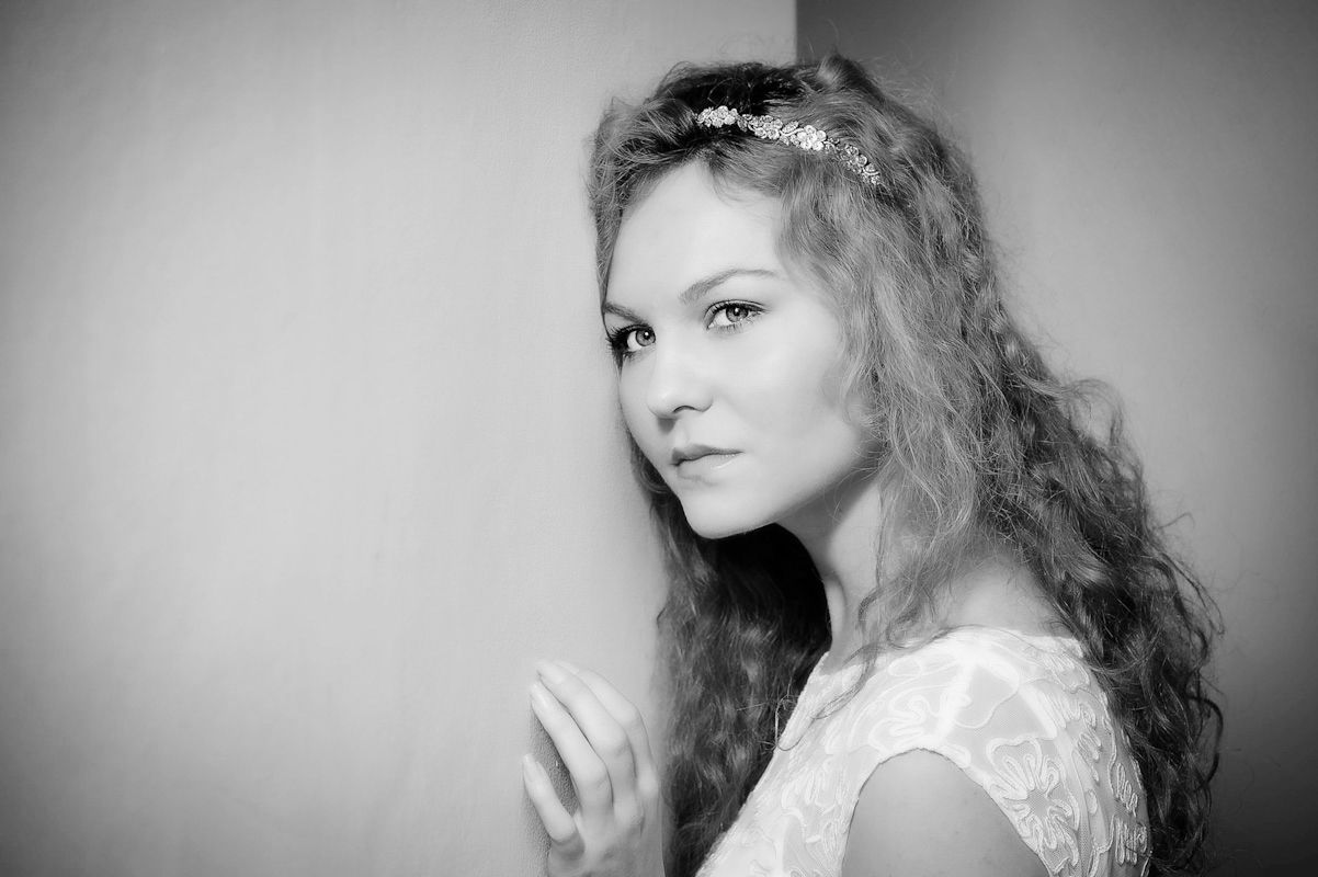 Black and white portrait of a young woman with curly hair wearing a white dress and floral headband leaning against a wall.