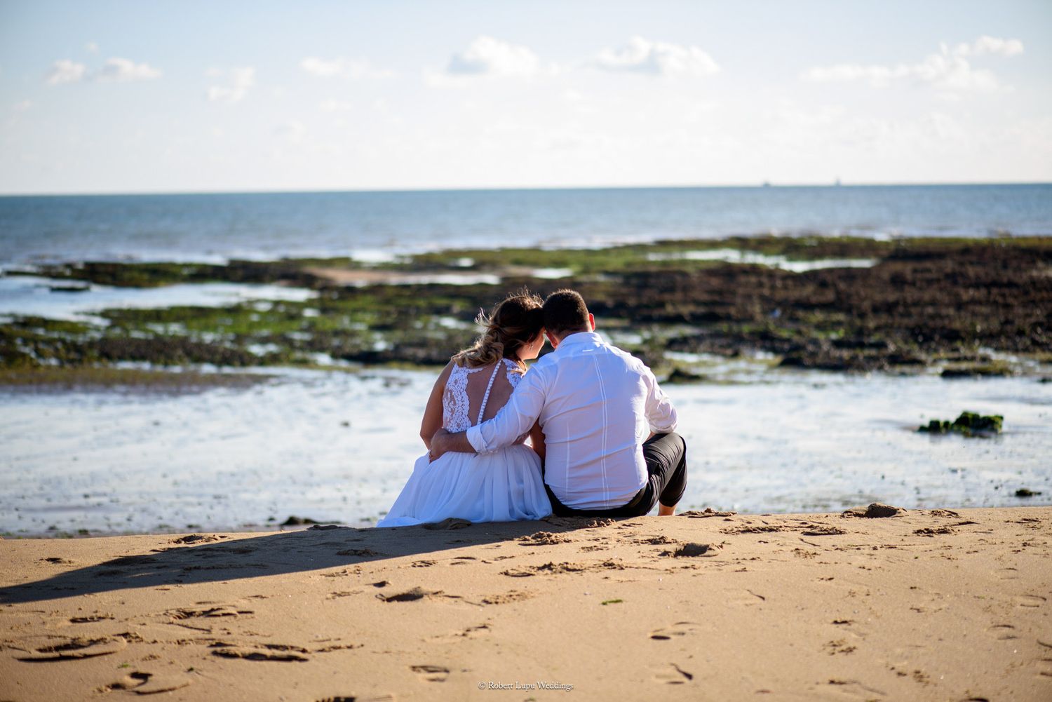 Newlywed couple sitting together on a sandy beach gazing at the calm ocean horizon.