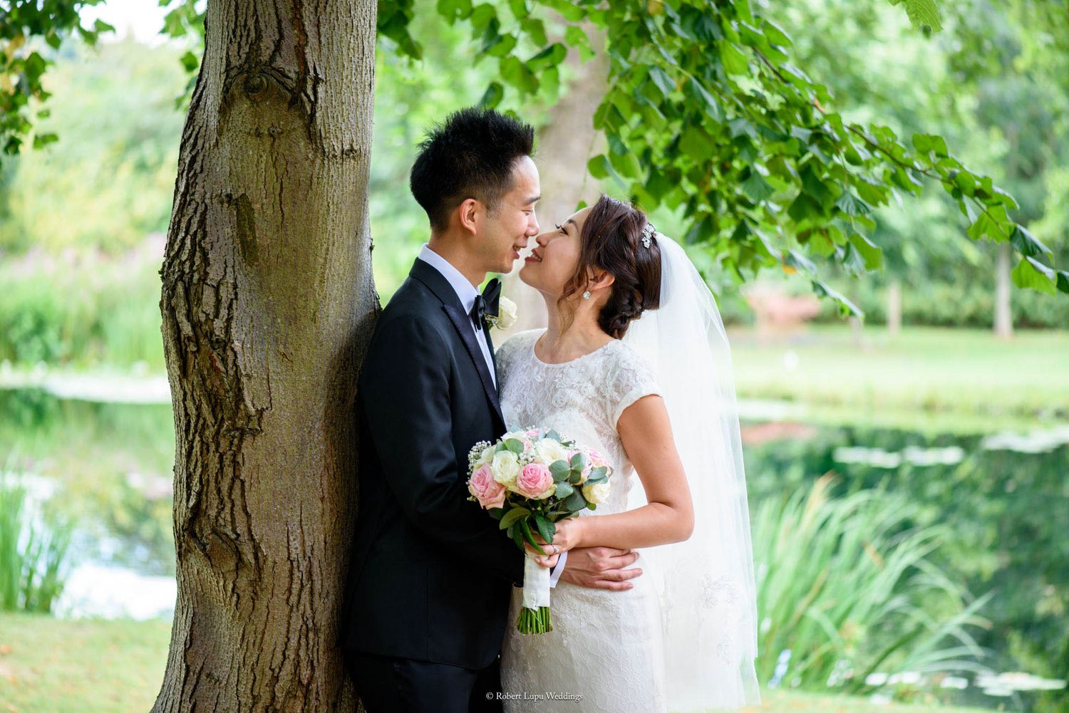 Asian couple in wedding attire sharing a romantic moment under a tree by a pond, bride holding a pink bouquet.