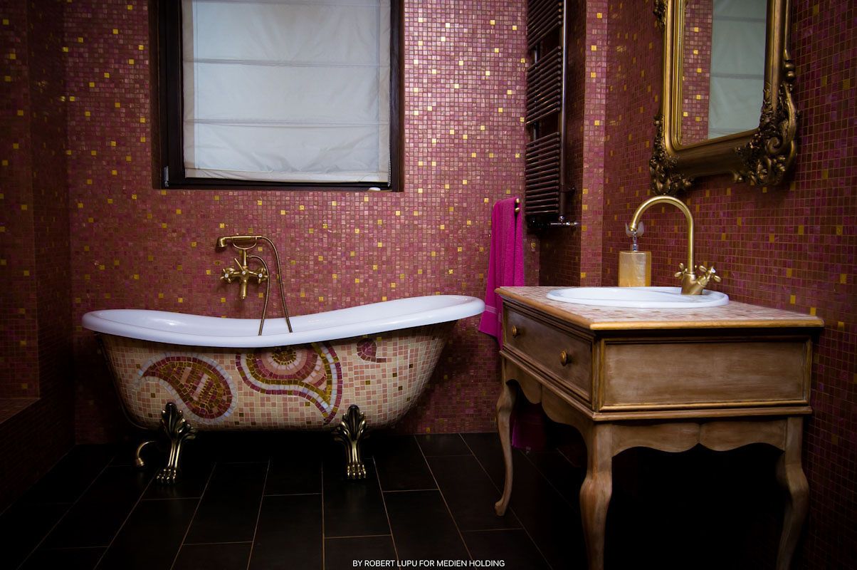 Ornate bathroom with clawfoot tub, mosaic tile walls in pink, gold fixtures, and a wooden vanity with mirror.