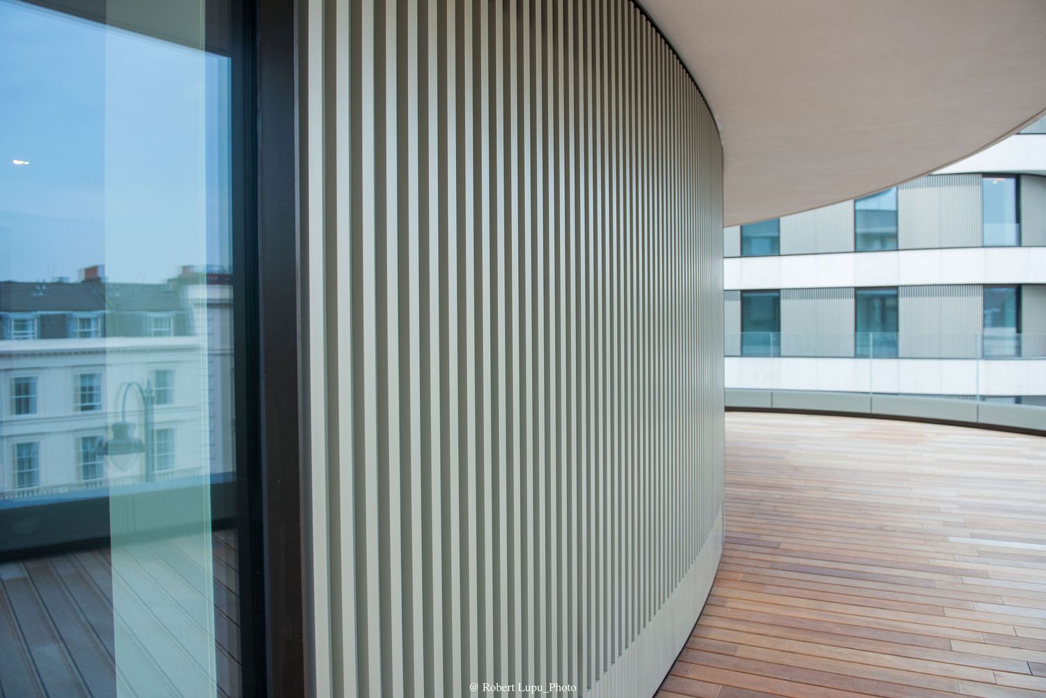 Modern balcony with curved vertical louvre screens, wooden decking, and glass railings overlooking a city building.