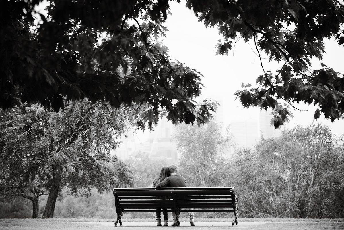 Couple sitting together on a park bench under leafy trees in a serene black and white landscape photo.
