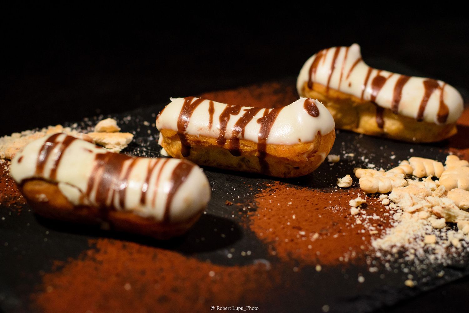 Chocolate drizzled eclairs with cream filling on a dark slate board dusted with cocoa powder.