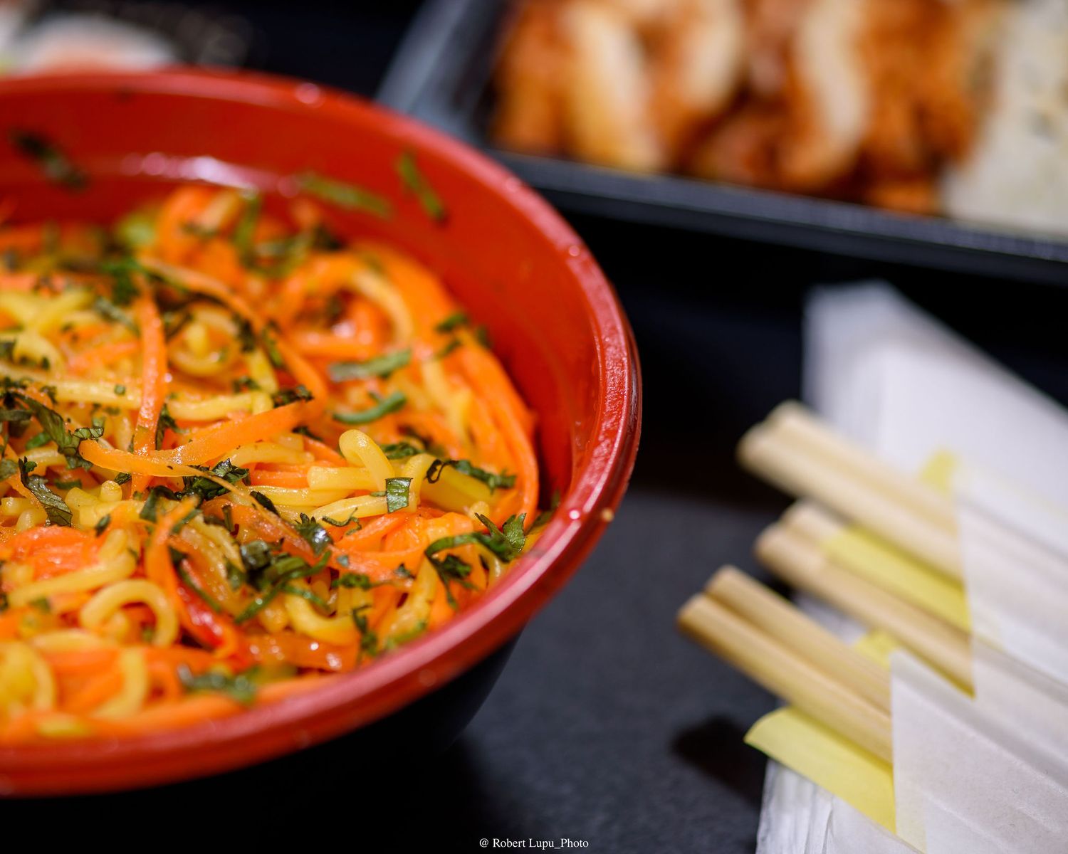 A vibrant red bowl filled with spiralized carrot noodles garnished with fresh herbs, served alongside lemongrass stalks.
