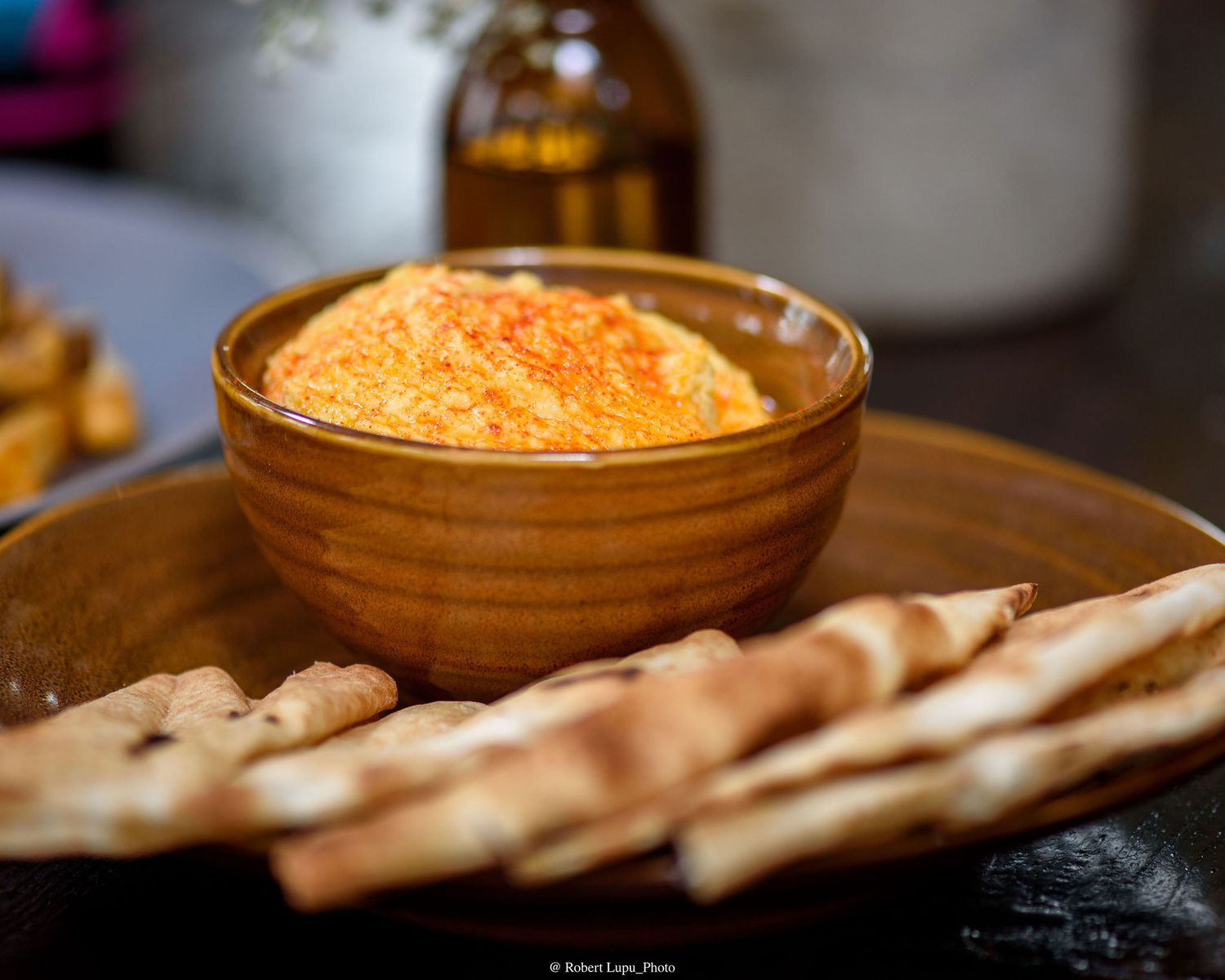 A bowl of creamy roasted red pepper hummus topped with paprika, served with crispy pita chips on a wooden plate.