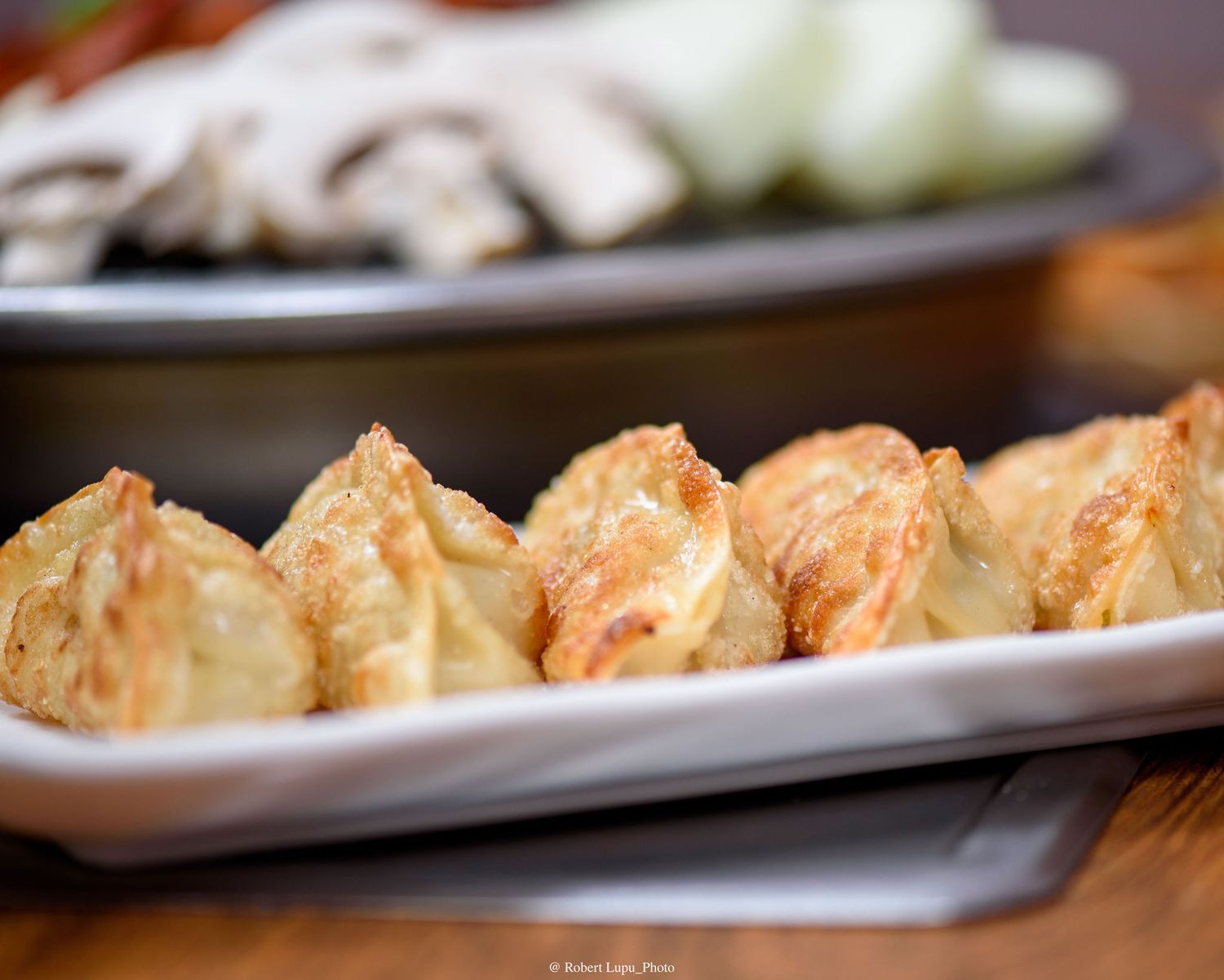 Golden pan-fried dumplings served on a white rectangular plate with mushrooms and onions in the background.