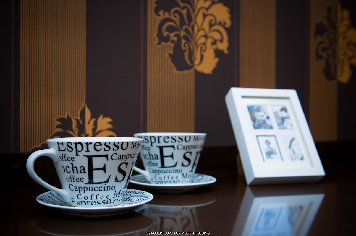 Large espresso-themed coffee cups with saucers displayed on shelf with framed photos in background.