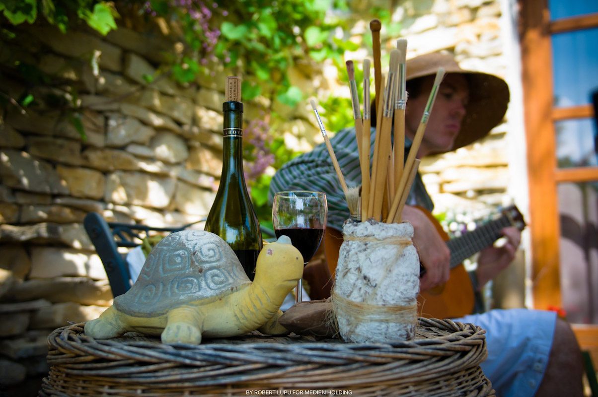 Rustic outdoor table with wine bottle, glass, breadsticks, and turtle figurine, with a person playing guitar in background.