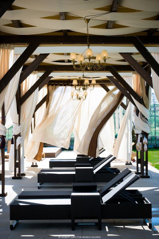 Elegant open-air pavilion with dark wooden beams, flowing white drapes, chandelier, and black lounge chairs in bright sunlight.