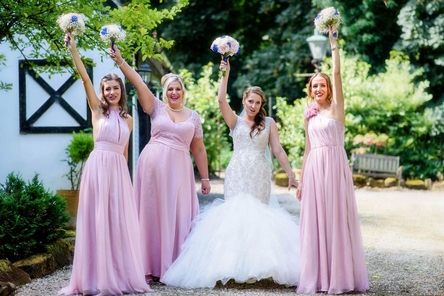 Bride in white gown celebrates with three bridesmaids in pink dresses, all raising bouquets outdoors.