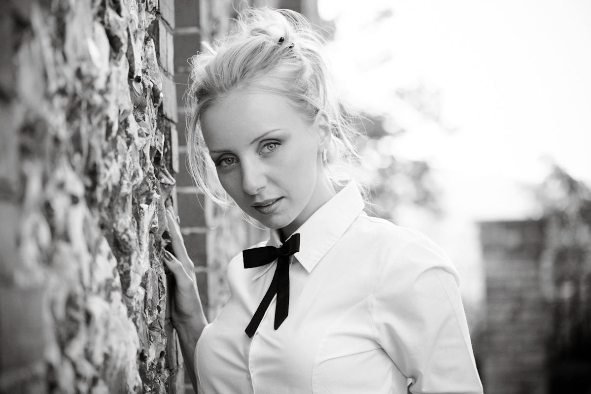 Black and white portrait of a blonde woman in a white shirt with a black bow tie leaning against a brick wall.