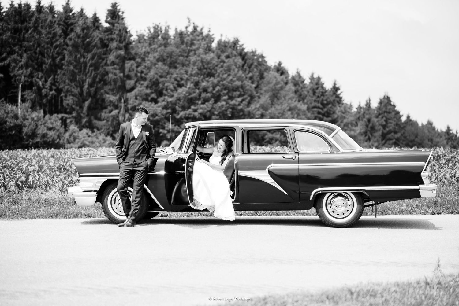 Black and white wedding photo of bride and groom posing with a classic vintage car on a sunny day.