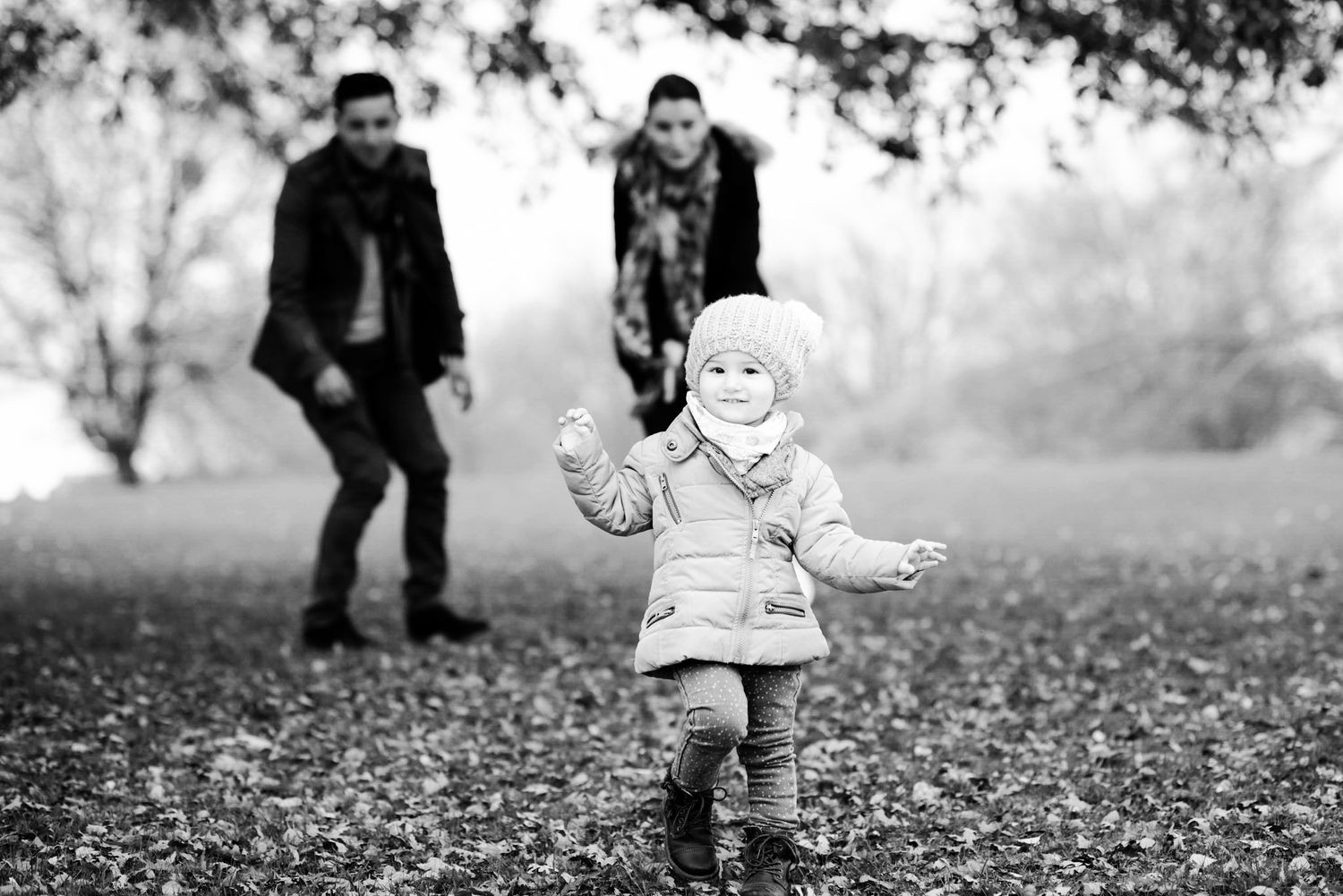 Black and white photo of a toddler walking in a park while two adults watch in the background.