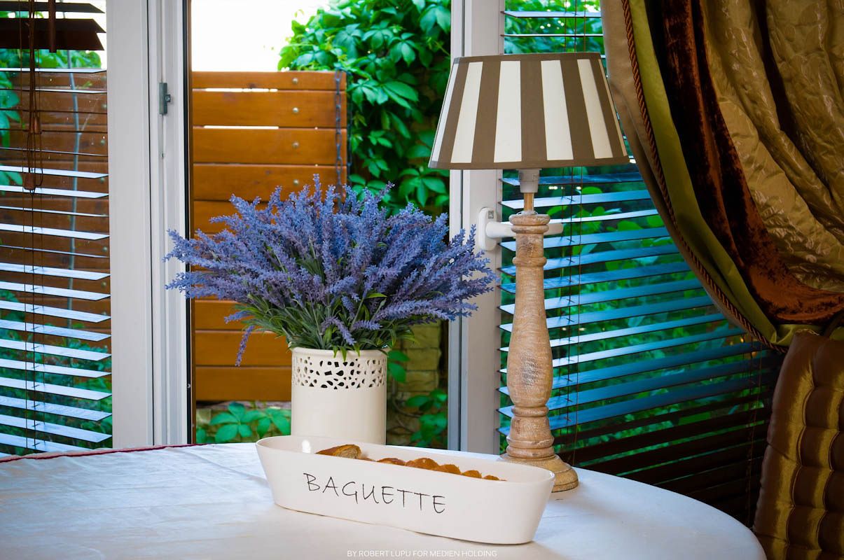 Elegant table with lavender flowers in a white vase, striped lamp, and a baguette tray near a sunlit window.