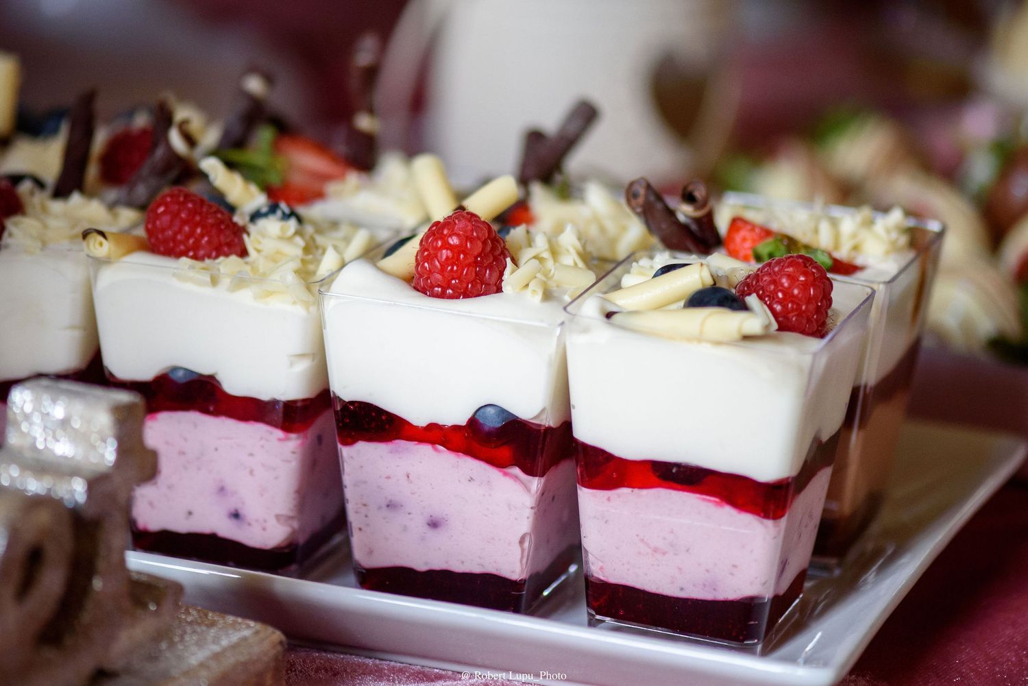 Elegant layered dessert cups with strawberry mousse, berry jam, white cream, fresh raspberries, and chocolate shavings on a white plate.