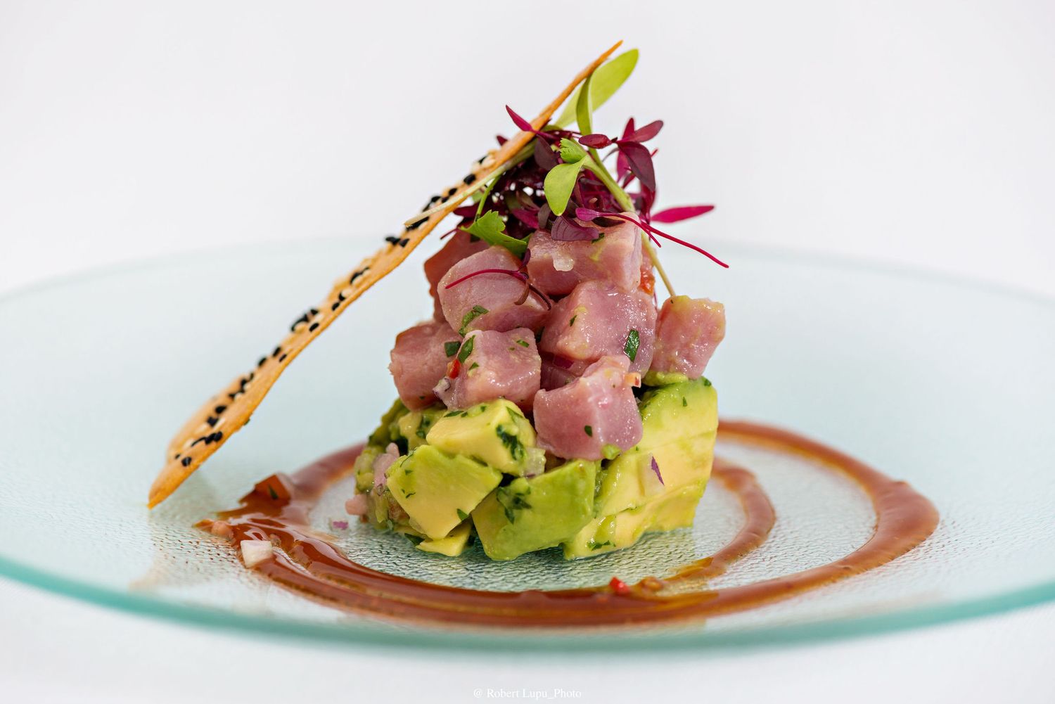 Elegant tuna tartare tower with avocado, microgreens, and sesame crisp on a white plate with sauce.