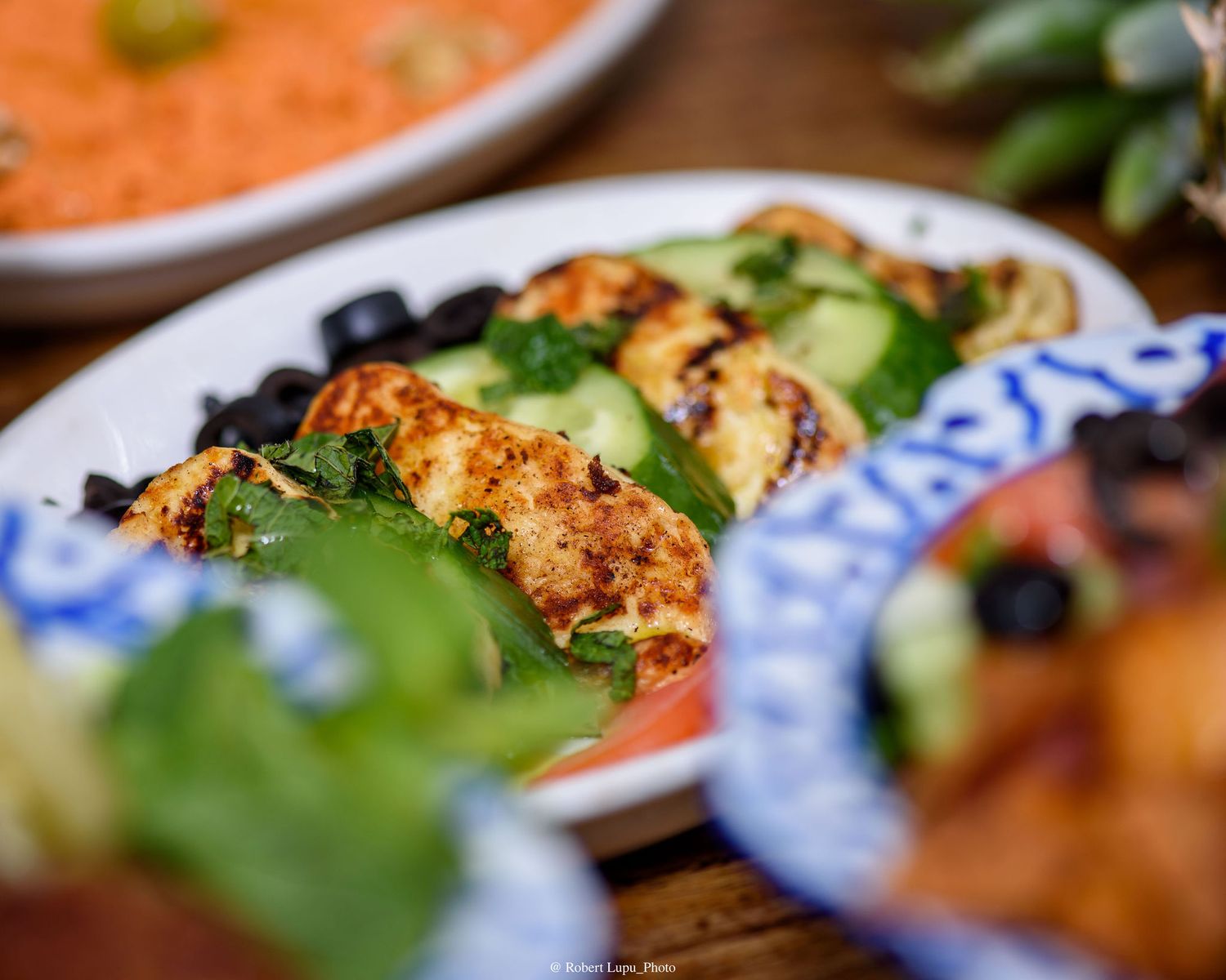 Grilled halloumi with cucumber slices, olives, and fresh greens on a decorative blue and white plate.