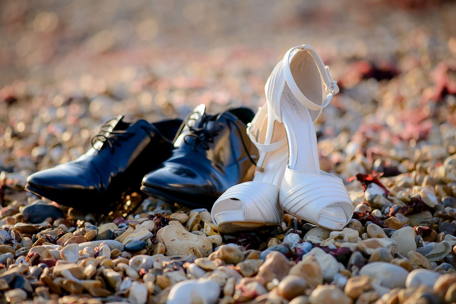 Black dress shoes and white bridal heels resting on a pebble beach, symbolizing a romantic wedding day.