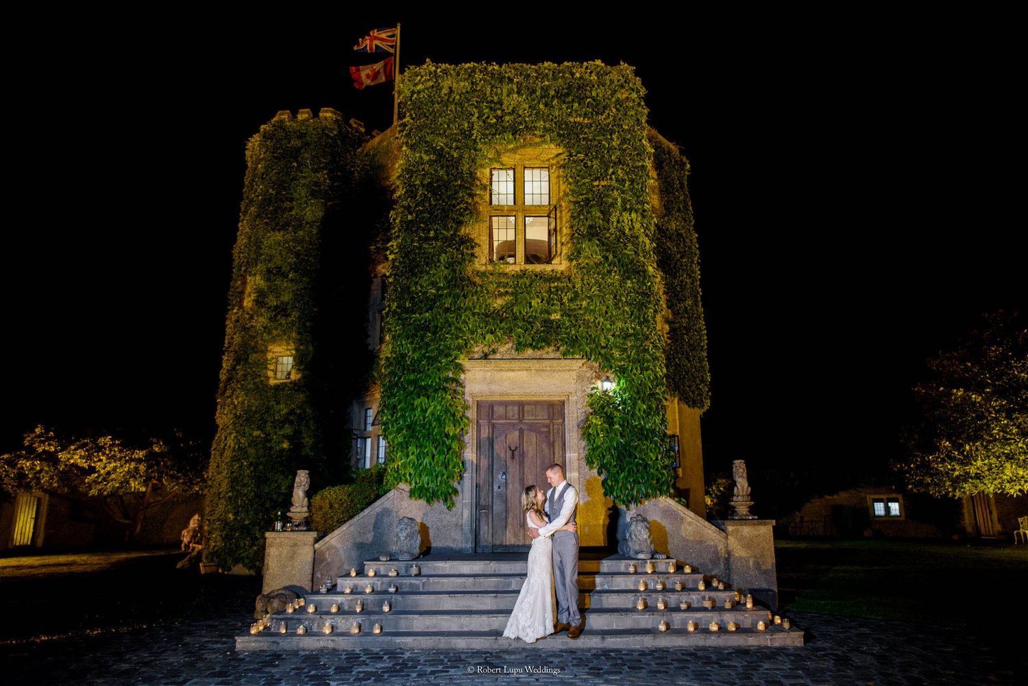 Bride and groom embrace on candlelit steps of ivy-covered castle at night during romantic wedding celebration.