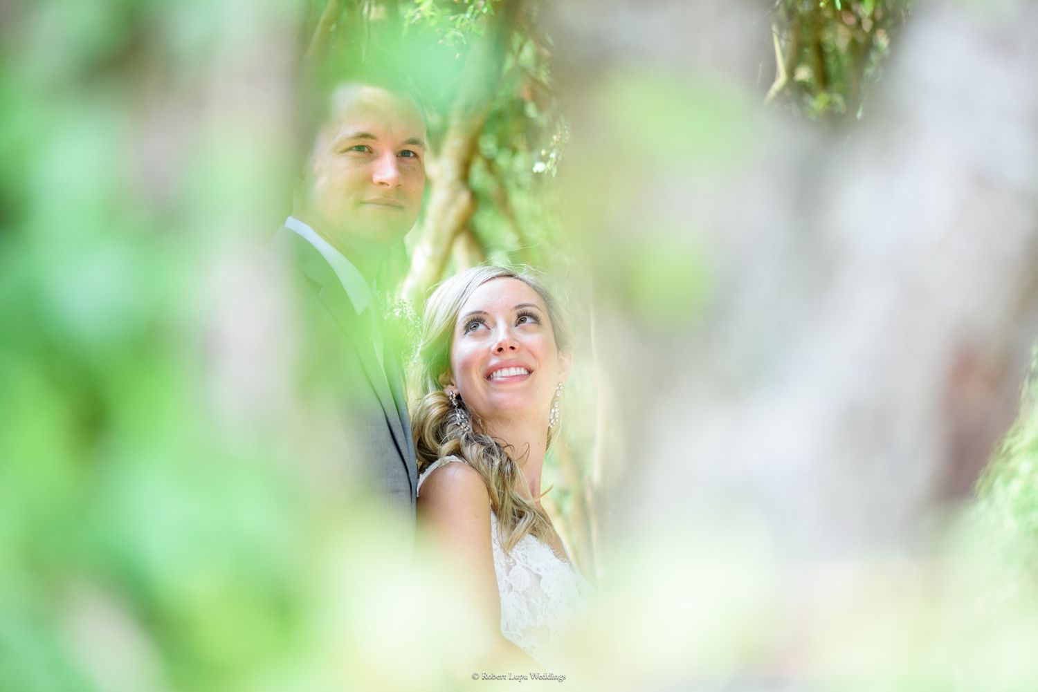 Bride and groom share a romantic moment framed by lush green foliage, bride gazing upward with a joyful smile.