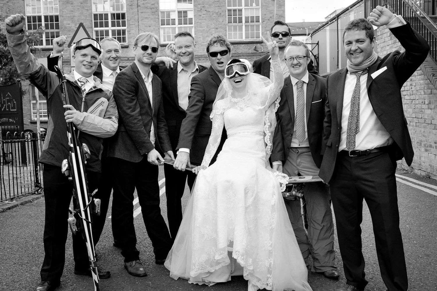 Black and white wedding photo of bride in white gown posing with groomsmen celebrating outdoors near brick building.