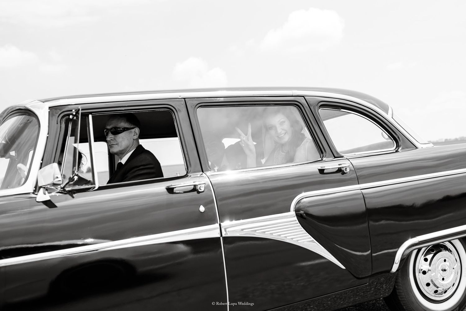 Black and white photo of a man in a cap driving a classic vintage American car on an open road.