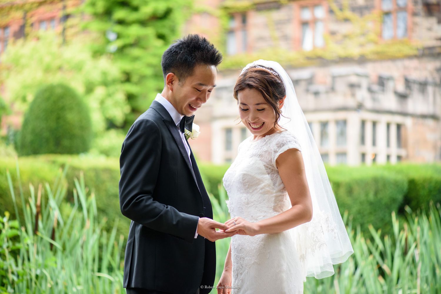 Asian bride and groom exchanging rings outdoors at elegant manor wedding venue surrounded by lush greenery.