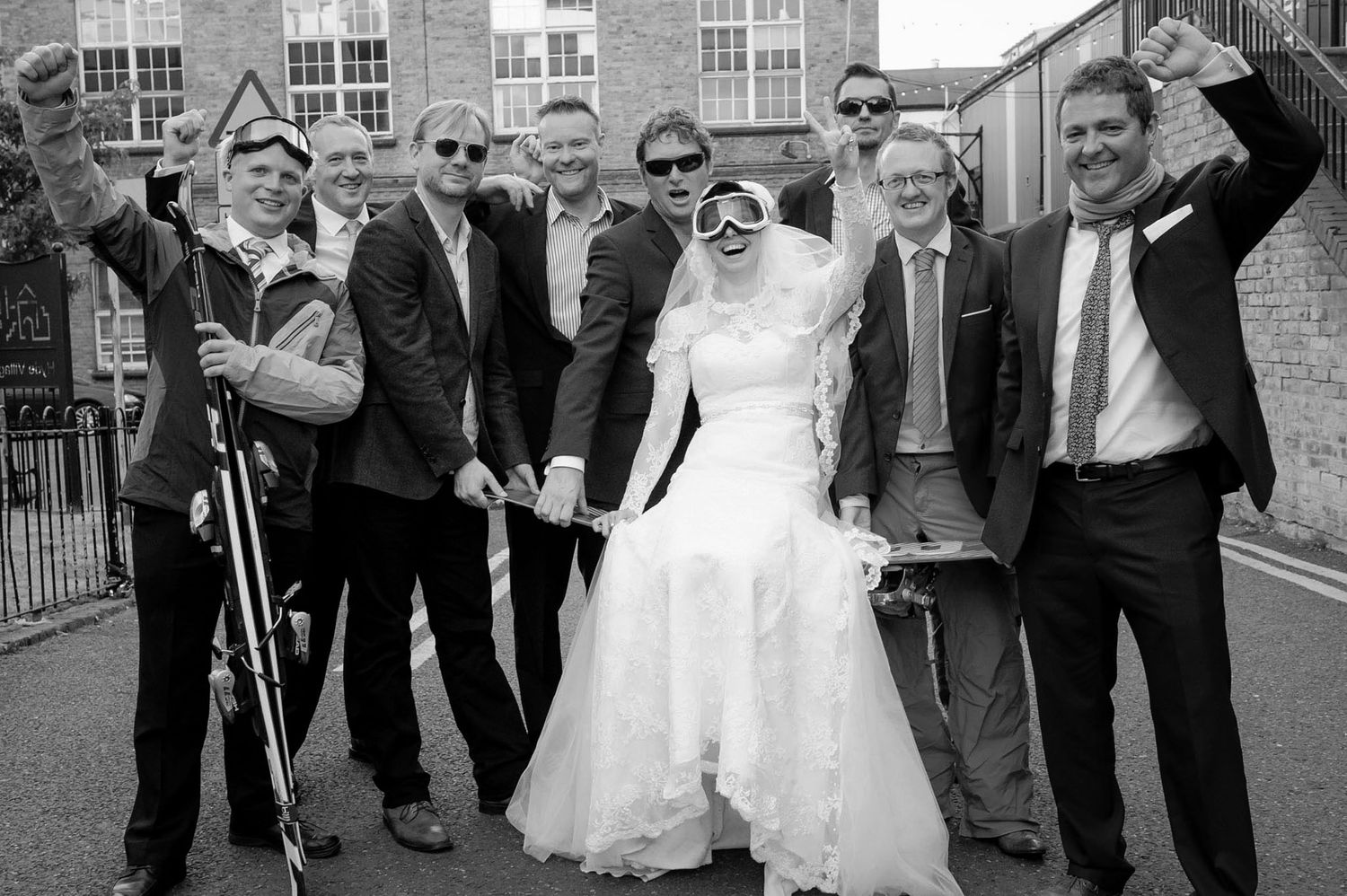 Black and white wedding photo of bride in white gown posing with groomsmen celebrating outdoors.