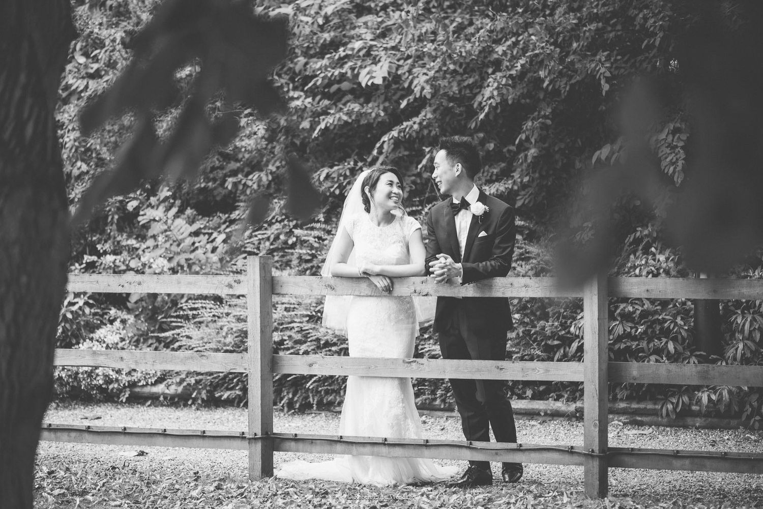 Black and white wedding photo of bride and groom smiling at each other by a wooden fence surrounded by lush trees.