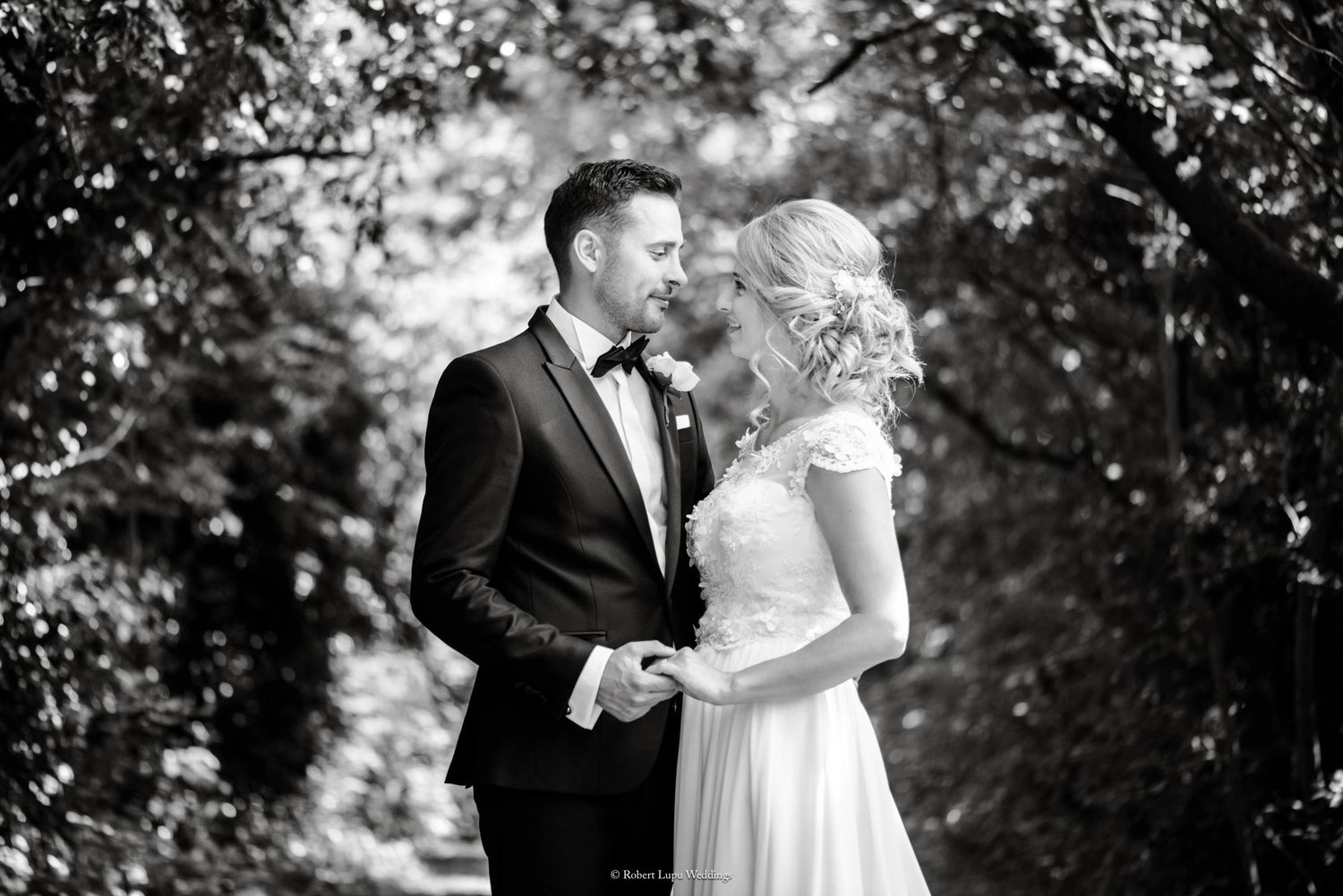 Black and white wedding photo of bride and groom gazing at each other surrounded by lush green trees.