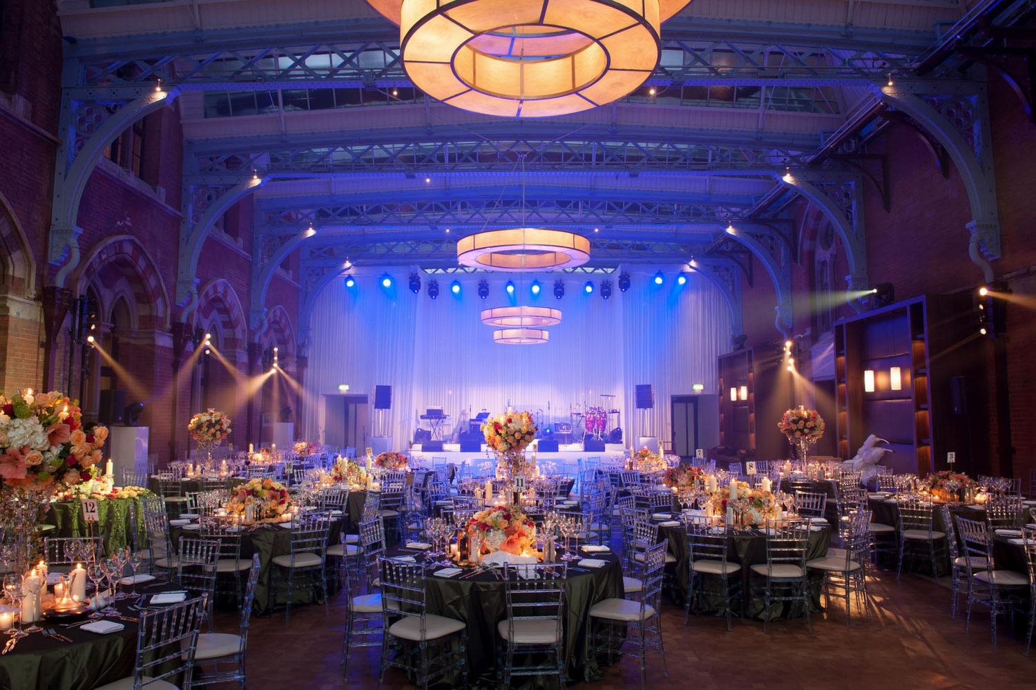 Elegant wedding reception hall with round tables, chiavari chairs, floral centerpieces, and colorful stage lighting in a grand ballroom.