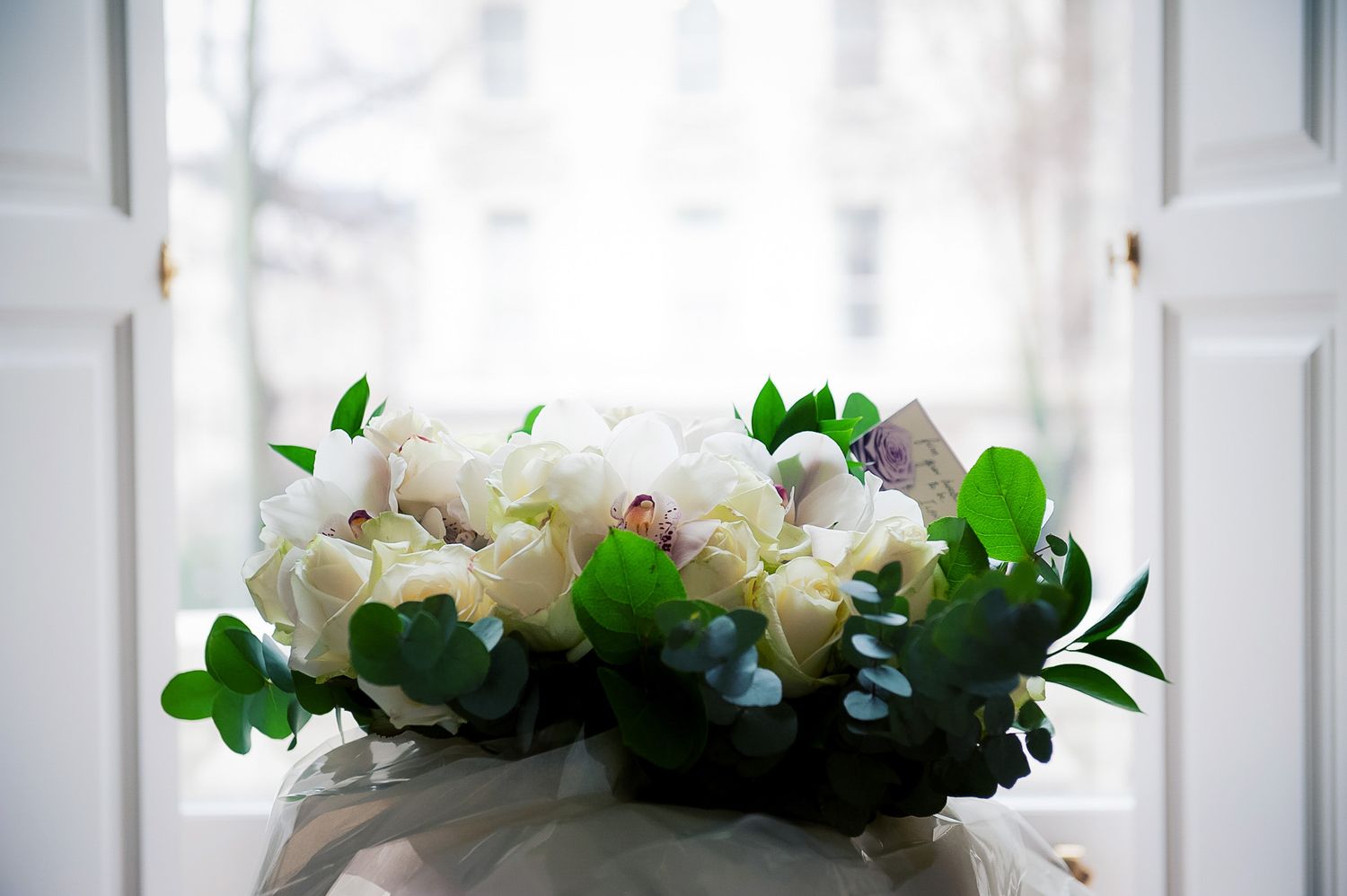 Elegant bridal bouquet of white roses and green leaves held near white doors.