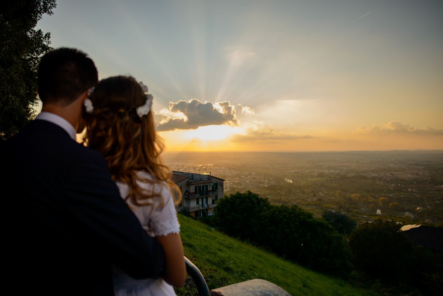 Bride and groom watching a stunning sunset over a scenic valley from a hilltop on their wedding day.