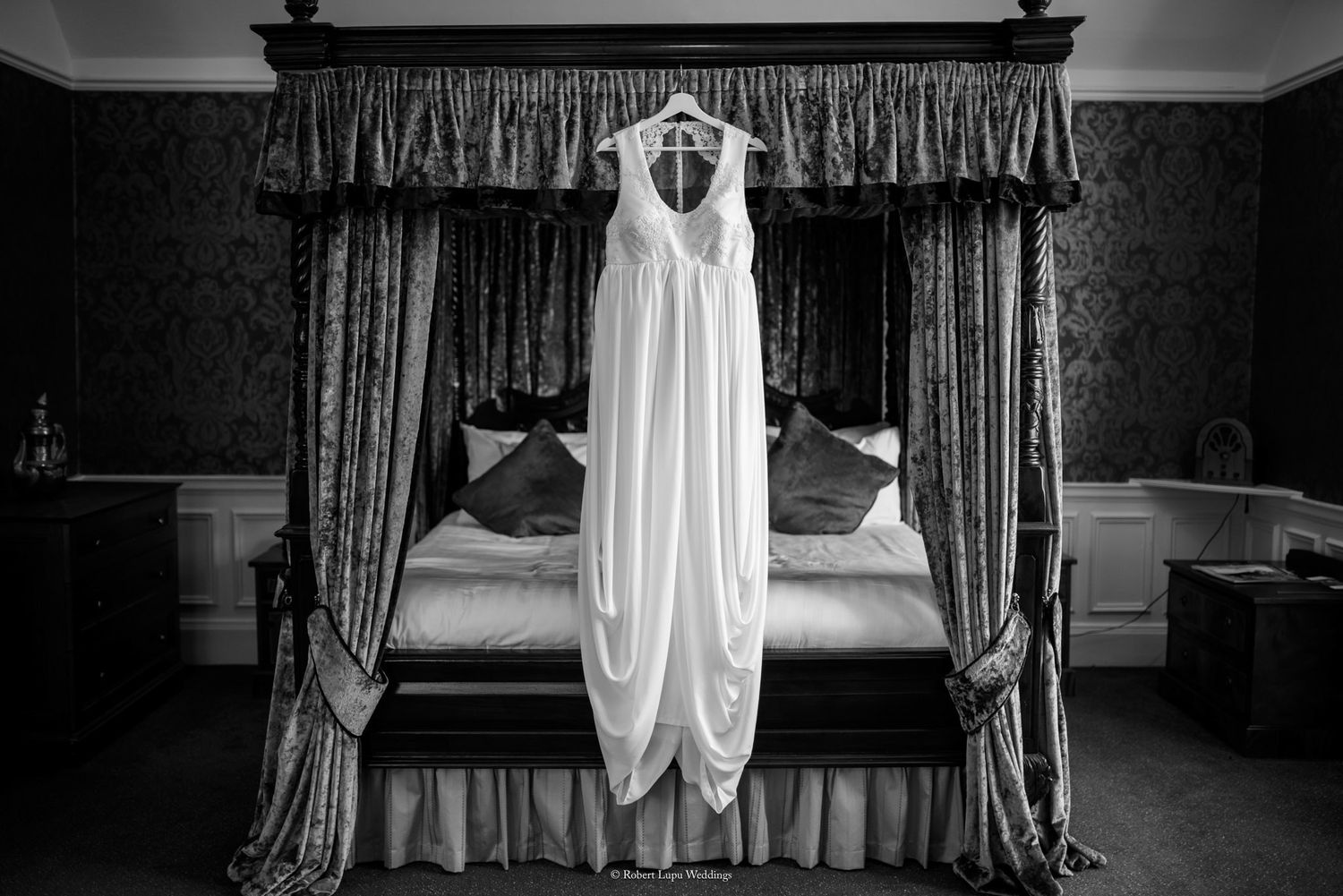 Elegant white wedding dress hanging on a ornate four-poster bed in black and white.