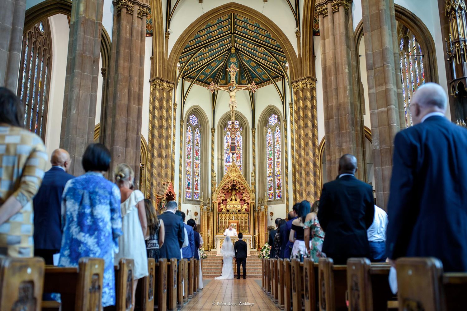 Wedding ceremony in a grand church with stained glass windows, guests standing in pews as couple exchanges vows at altar.