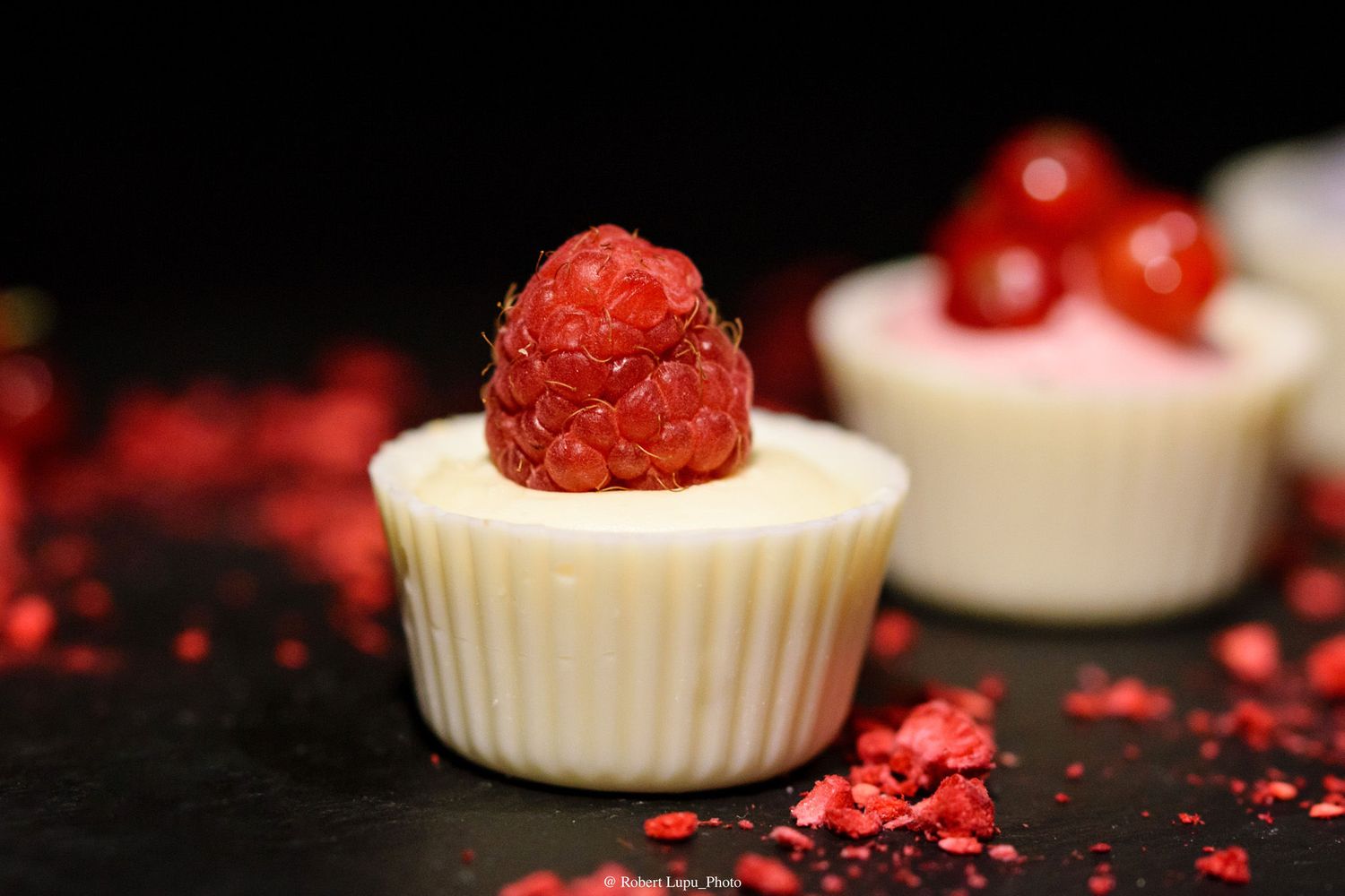 White chocolate mini cupcakes topped with fresh raspberries and red currants on a dark surface.