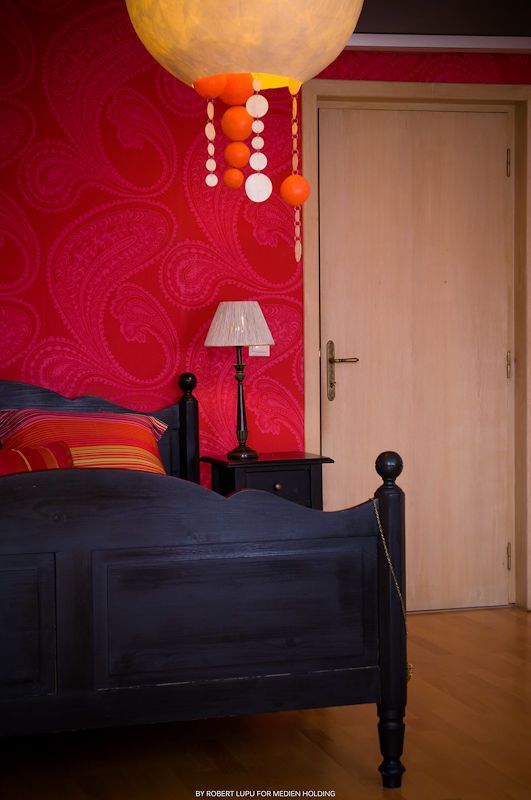 Cozy bedroom with bold red paisley wallpaper, black wooden bed, orange pendant lamp, and bedside table lamp.