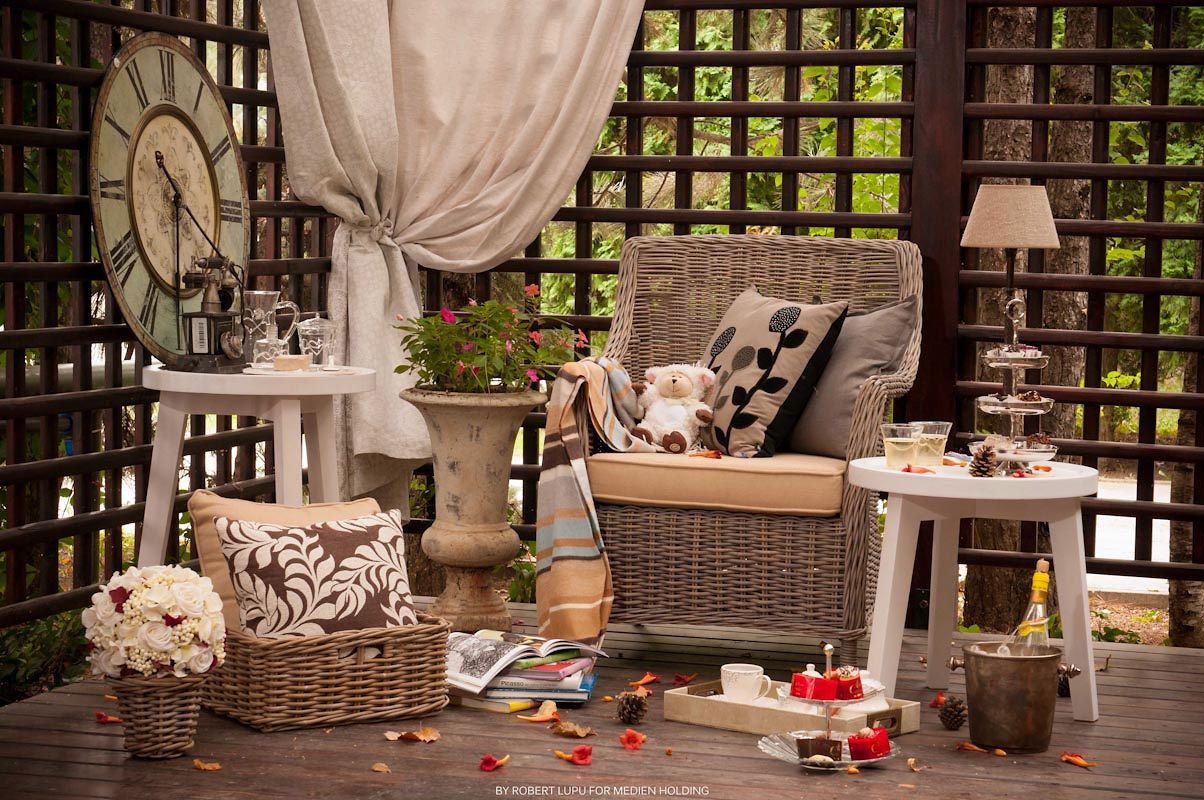 Cozy wicker patio furniture with decorative pillows, clock, and floral accents on a dark wood pergola deck.