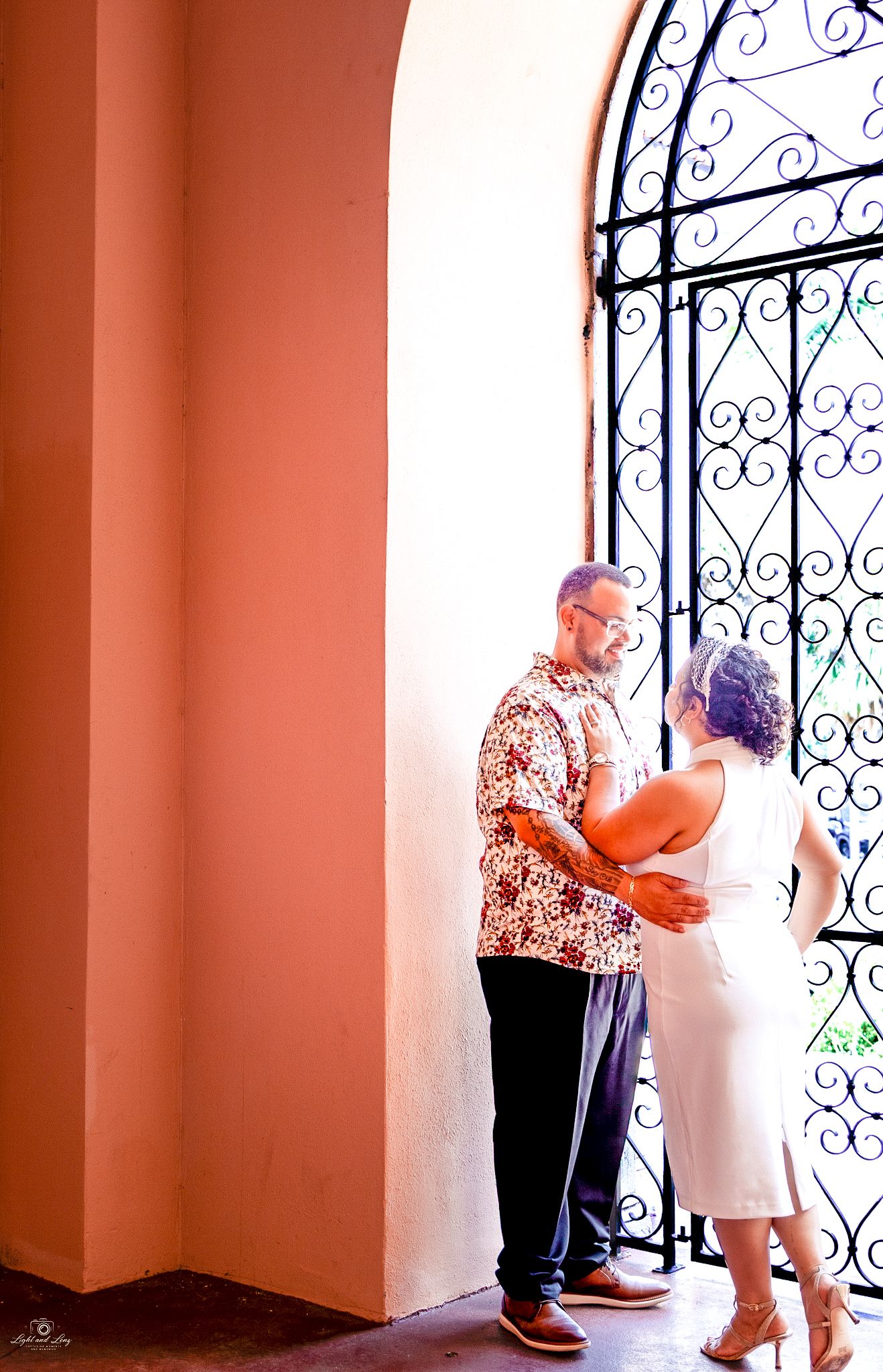 Wedding couple standing together near a wrought iron arched window in a warm terracotta building.