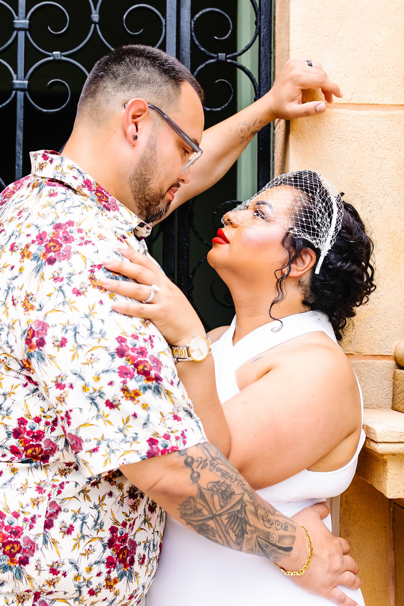 Groom in floral shirt leaning toward bride in white dress against ornate iron gate exterior wall.