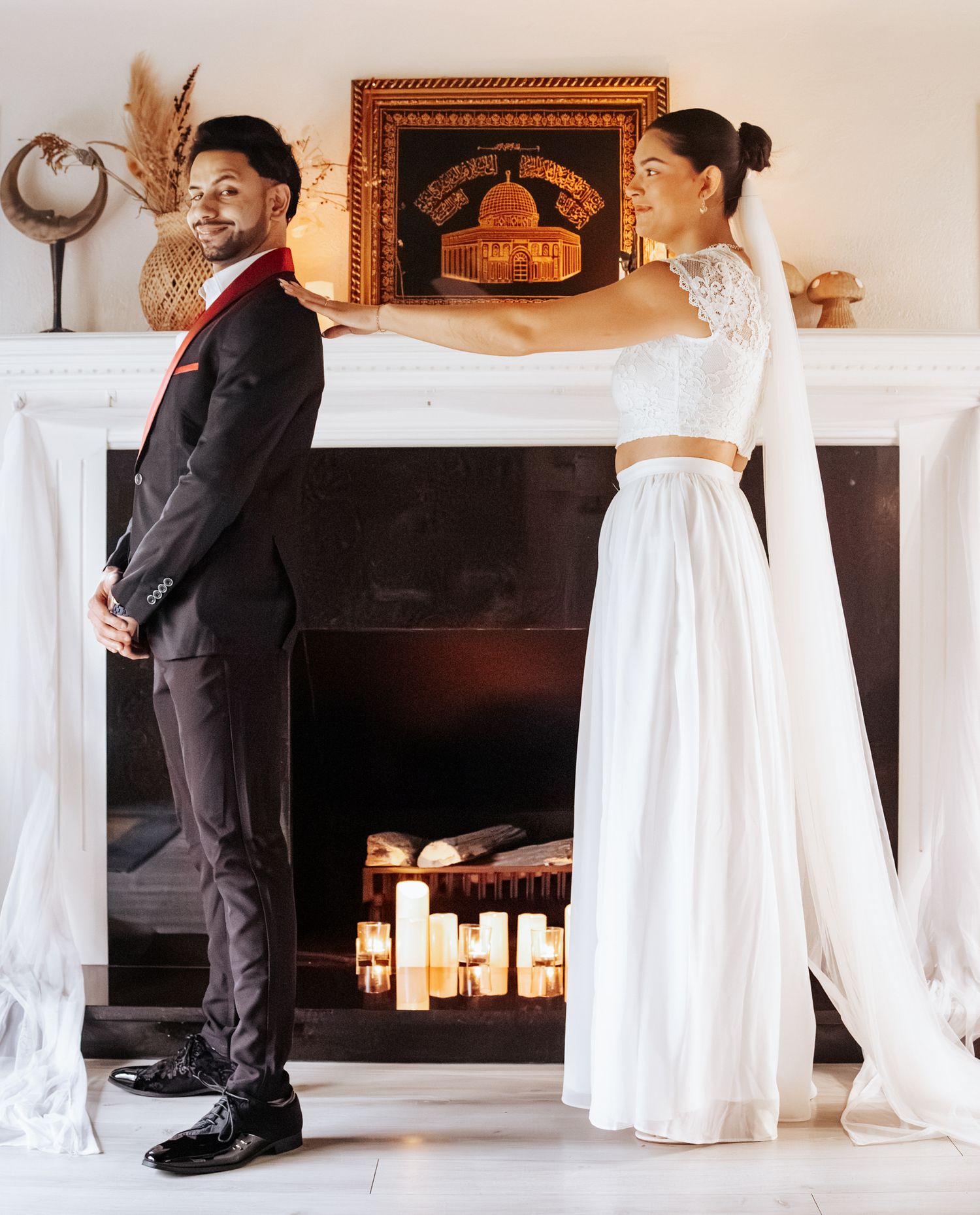 Bride in a white two-piece gown and long veil reaches toward a groom in a dark suit as they stand before a lit fireplace.