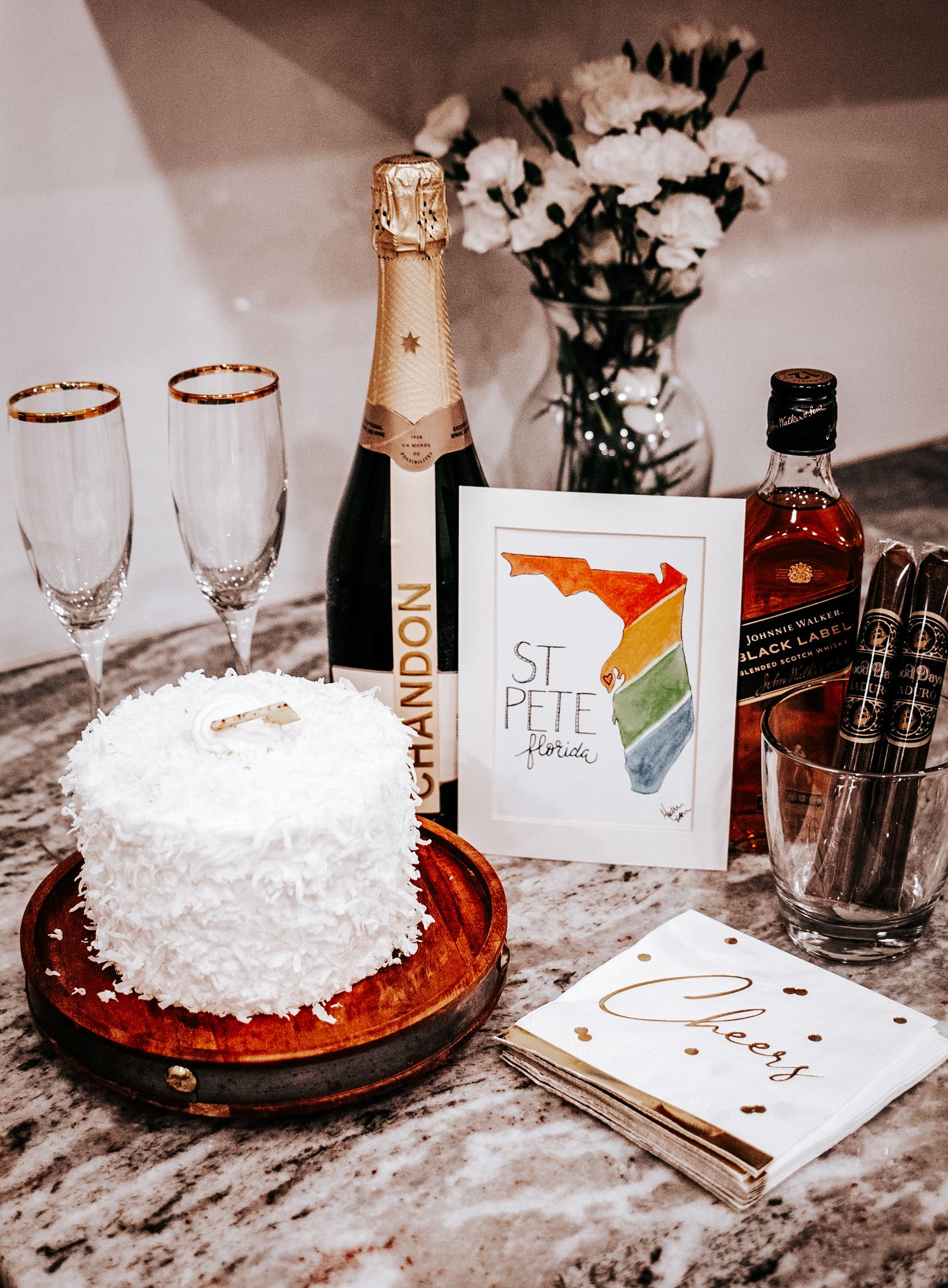White coconut cake beside Chandon champagne, two flutes, a rainbow St Pete Florida card, whiskey, cigars and a Cheers napkin on granite.
