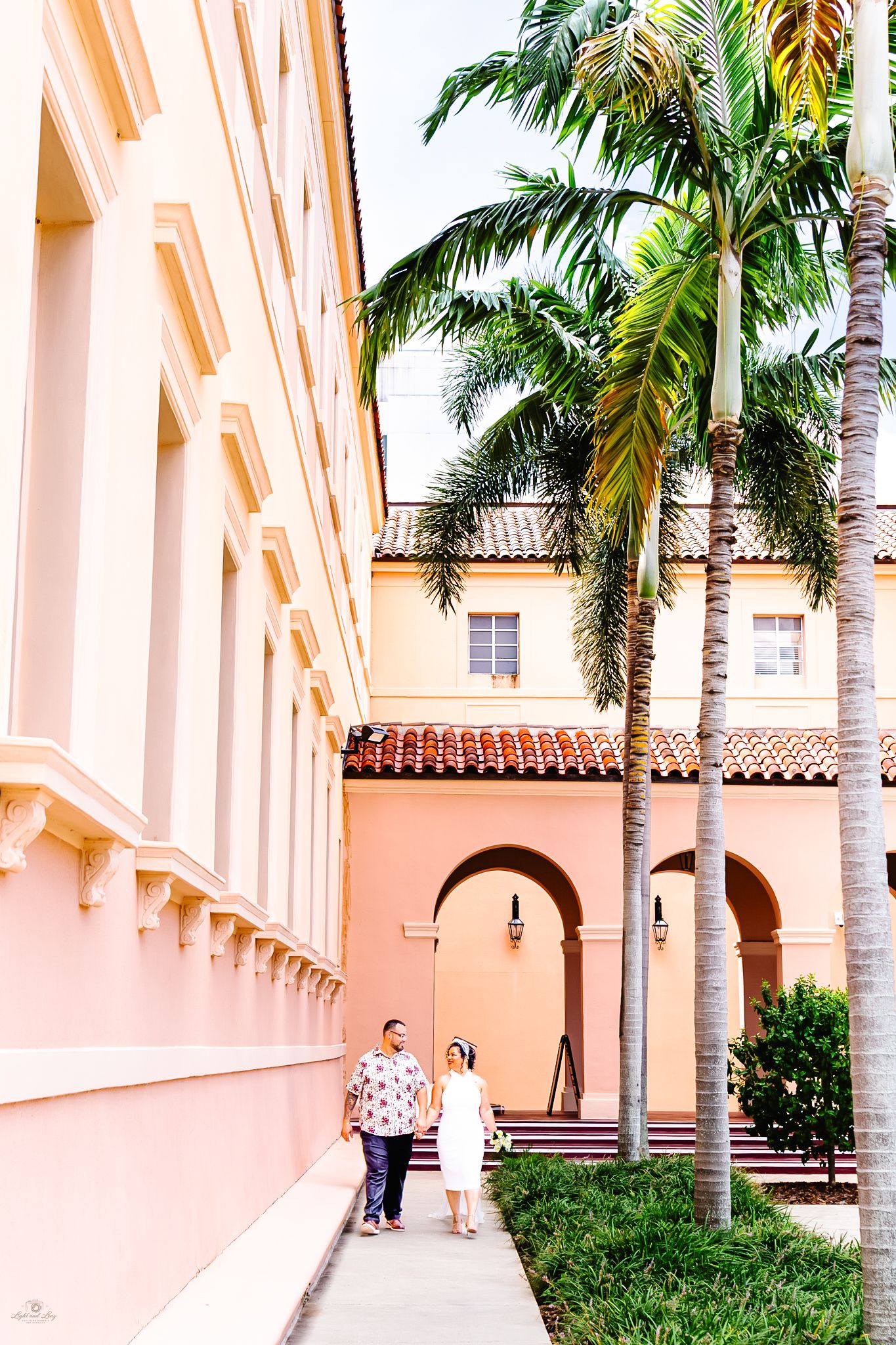 Newlyweds walking hand in hand along a pastel pink building exterior lined with tall palm trees.