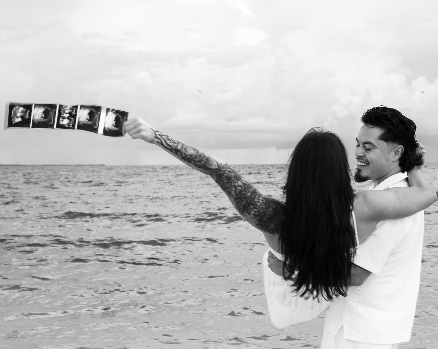 Couple embracing on a beach in black and white as the woman with long hair and a tattooed arm holds up a strip of ultrasound photos.