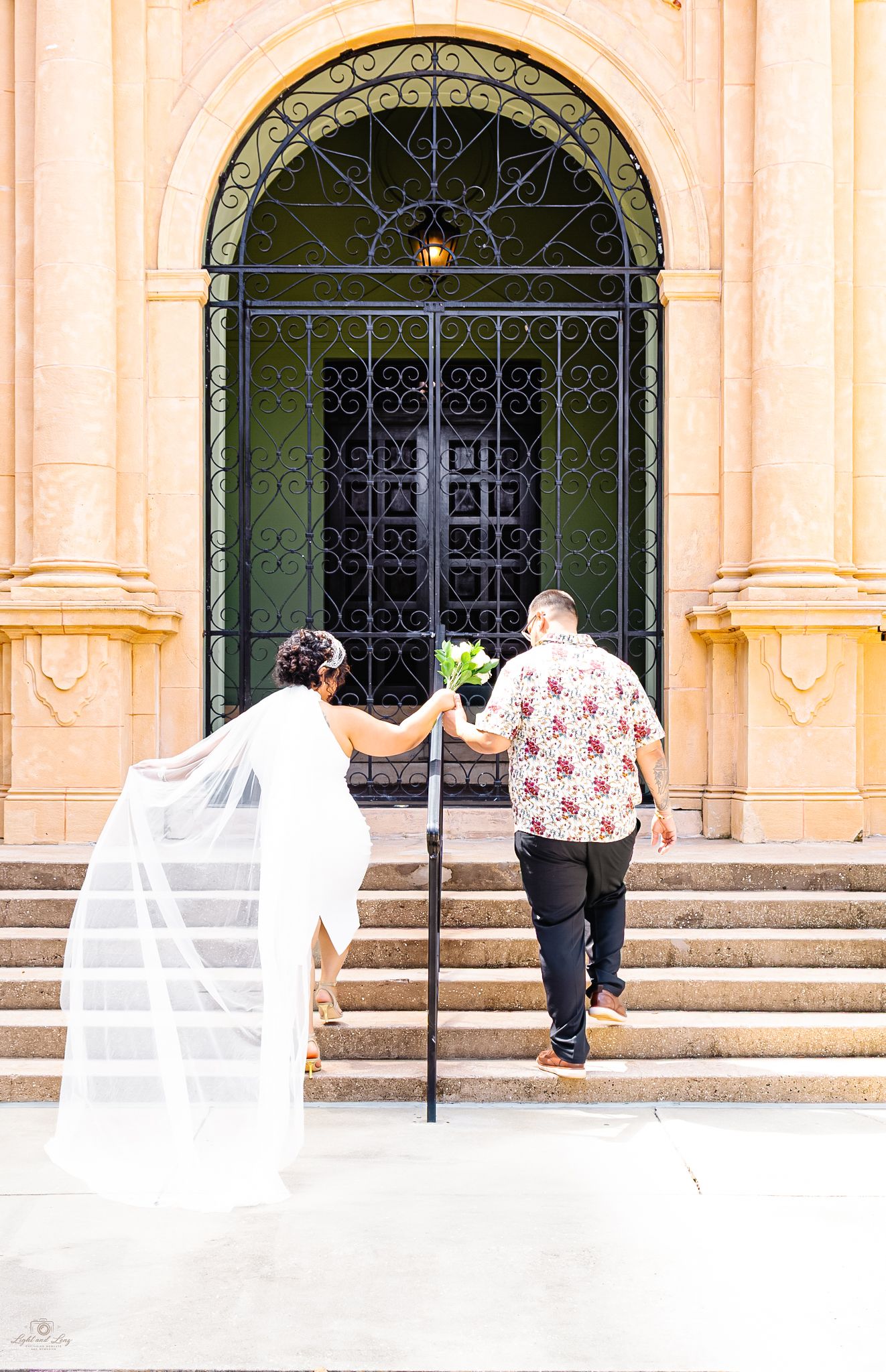 Bride in white gown and veil holds bouquet with groom ascending steps to ornate iron gate of elegant venue.