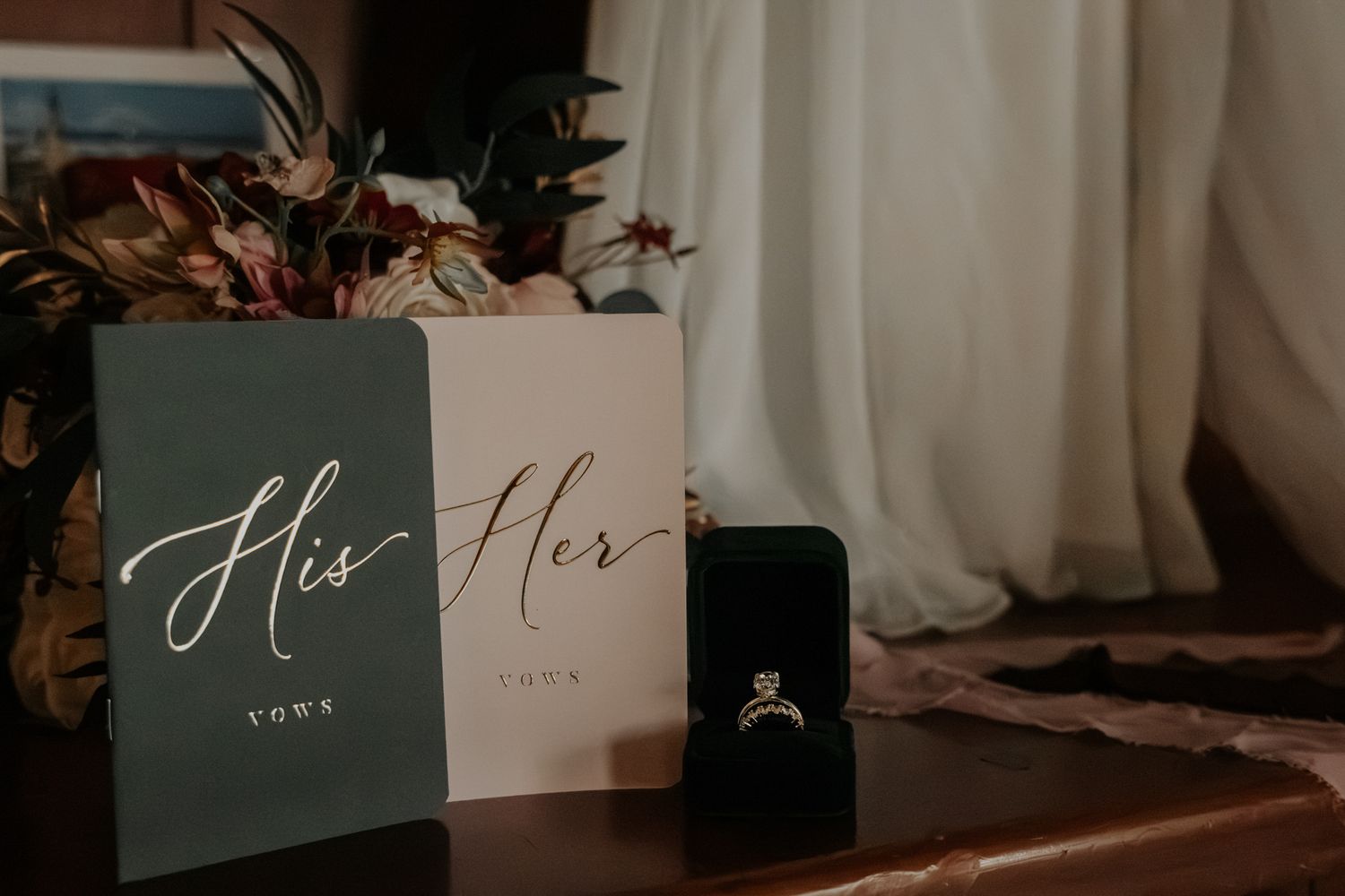 Green His and cream Her wedding vow books beside a diamond engagement ring in a black box, with dried floral decor and soft curtains.